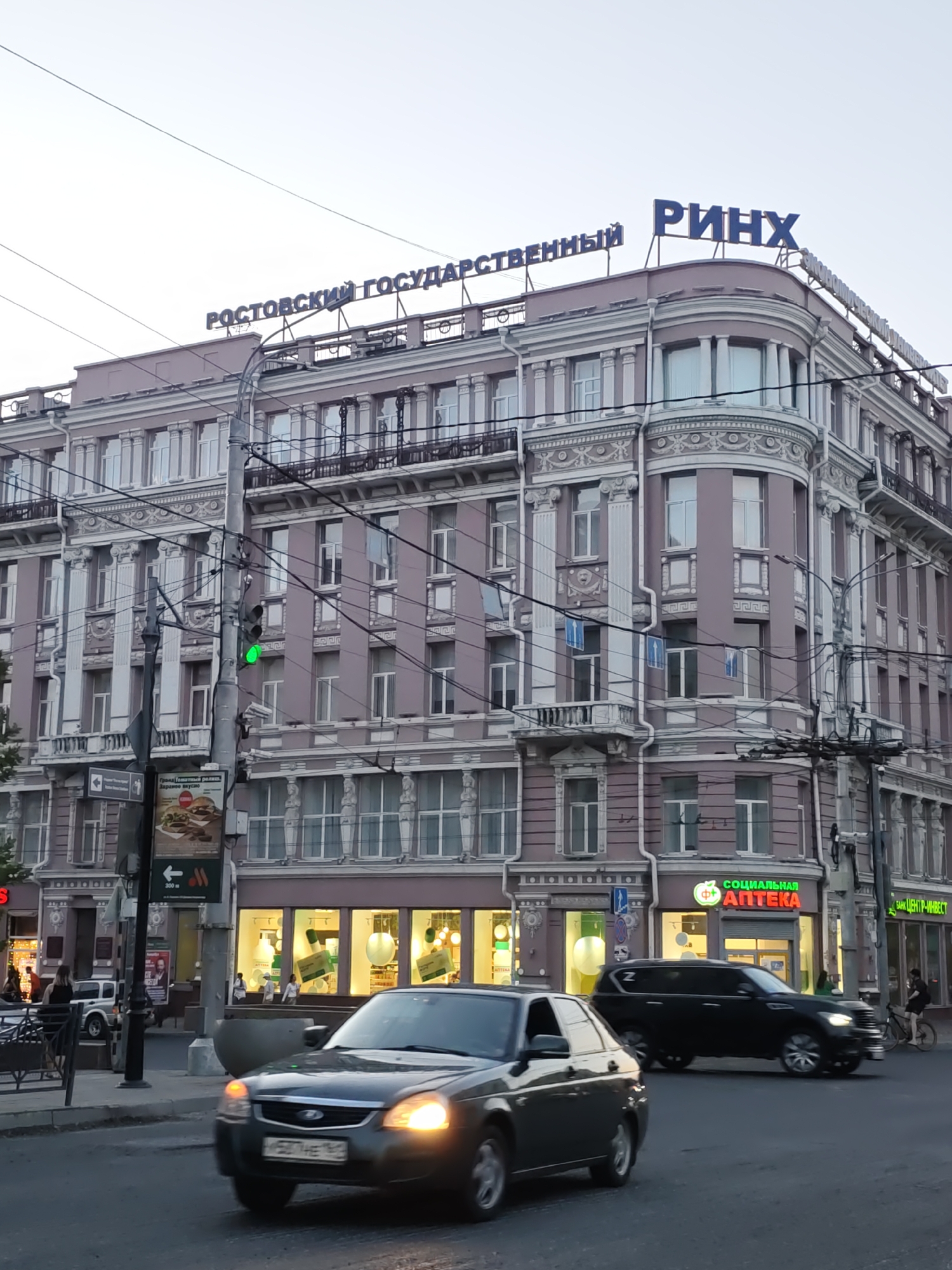 The image shows a street scene in Rostov-on-Don, Russia, with a large, ornate building dominating the frame. The building features multiple stories, decorative architectural elements, and large windows. On the rooftop of the building, there are prominent blue signs with Cyrillic text. The left sign reads "РОСТОВСКИЙ ГОСУДАРСТВЕННЫЙ" and the right sign reads "РИНХ". These likely refer to the Rostov State University of Economics (RSUE), also known as RINKh.
In the foreground, two cars are visible on the street. A dark sedan is in motion on the left, and a black SUV is to its right, also in motion. A traffic light with a green light illuminated is visible on a pole on the left side of the street.
Below the rooftop signs, on the lower levels of the building, there are storefronts. One storefront has a green and white sign that reads "СОЦИАЛЬНАЯ АПТЕКА" (Social Pharmacy). Another sign, further to the right, appears to say "БАНК ЦЕНТР-ИНВЕСТ".
There are several people visible in the scene, including individuals walking on the sidewalk and in front of the storefronts. The sky is overcast, suggesting it might be evening or a cloudy day. Overhead wires and poles are present, indicating a street with public transportation or utilities. The overall impression is a busy urban environment.