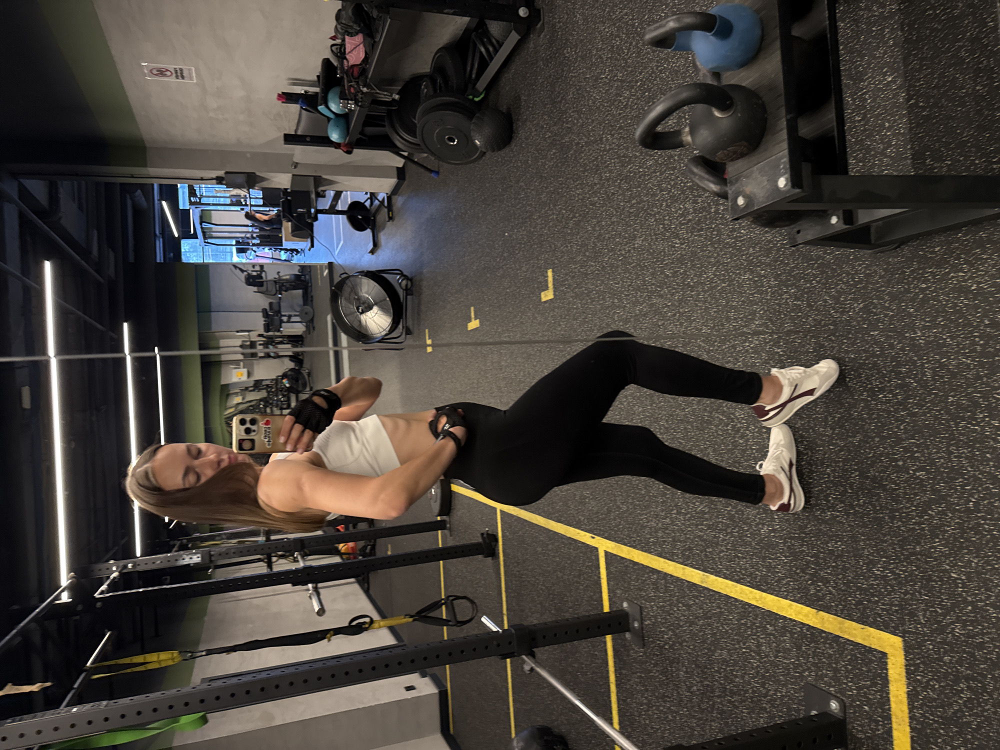 The media file depicts a woman in a gym, taking a selfie in a mirror. The woman is wearing a white sports bra and black leggings, with white athletic shoes. She has long, brown hair and is wearing black fingerless gloves. In the background, there is gym equipment, including a weight rack with kettlebells, weight plates, and a fan. The gym floor is dark grey with yellow markings. The lighting suggests it is likely indoors, possibly evening. The overall mood appears to be one of casual fitness. There is no visible text in the image. The location context is Sochi, Russia.