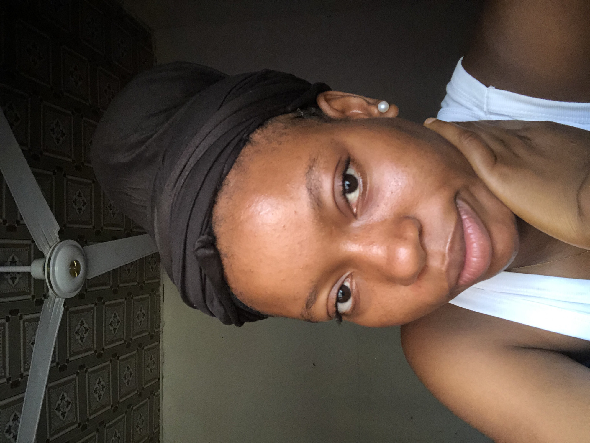 A woman is shown indoors in Kashere, Nigeria, likely in a residential setting. She has brown skin and a brown head covering, possibly a wrap. The woman is wearing a white top and a pearl earring. She is partially holding her neck with her hand. The background consists of a wall with patterned tiles and a ceiling fan. The lighting is somewhat dim.