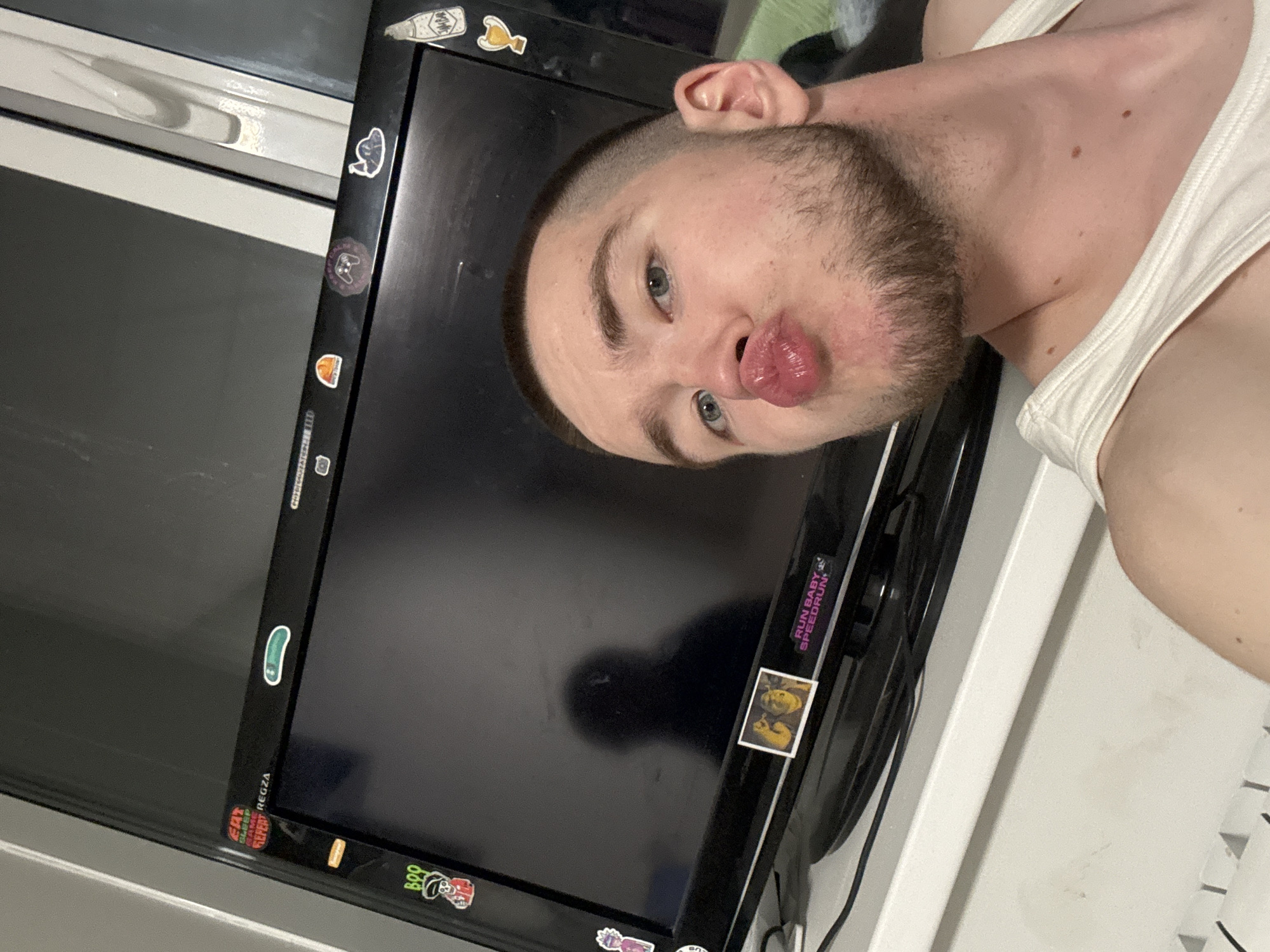 The image is a tilted selfie of a young man, presumably taken indoors in Kazan, Russia. The man has light skin, blue eyes, and a short beard, and is wearing a white tank top. His head is angled, and he is making a playful "fish face" or "duck lips" expression towards the camera.
He is positioned in front of a black computer monitor or television screen, which serves as a prominent background element. The monitor is adorned with various colorful stickers along its bezel. Visible text on these stickers includes "RUN BABY SPEEDRUN," "REGZA" (likely the monitor's brand), "EAT SLEEP REPEAT," and "BOO." Other stickers depict various graphics such as a bottle, a yellow duck or chicken, a ghost, a game controller, and a green bean or pickle shape. A clear reflection, possibly of the man's phone or a light source, is visible on the dark monitor screen.
The monitor rests on a white surface, possibly a desk or shelf, and a white window frame is visible in the background on the left side of the image, suggesting natural light. Wires are visible near the bottom right corner of the setup. The overall mood conveyed by the man's expression is lighthearted and humorous.