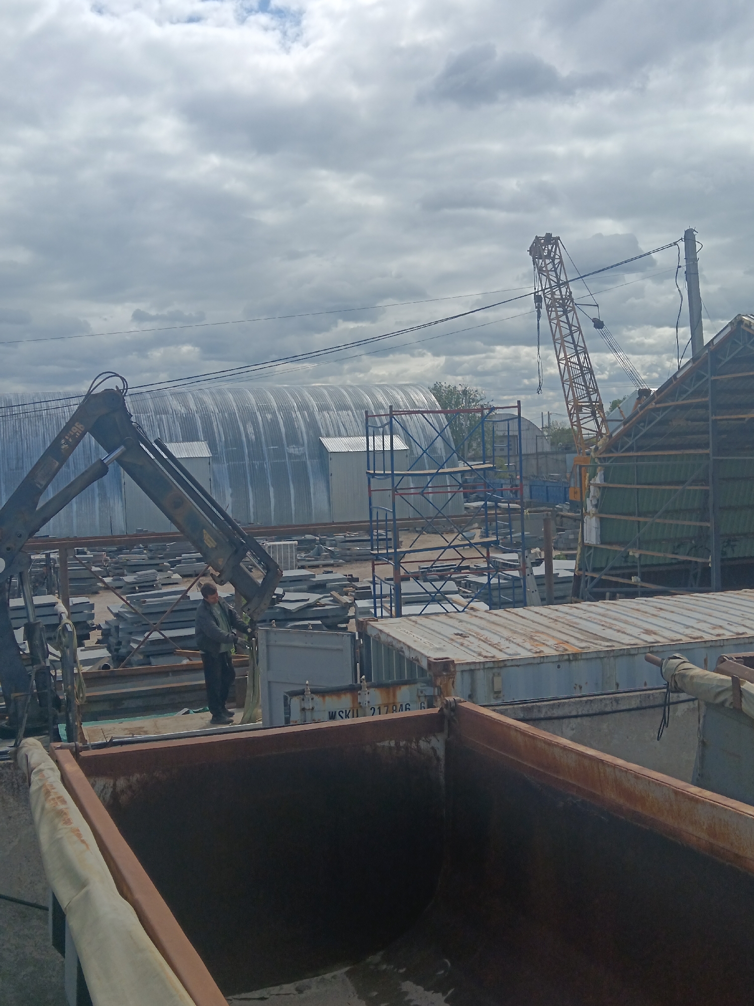 The image, taken in Яльгелево, Russia, shows a construction or industrial yard on an overcast day. A person wearing dark clothing stands on a flatbed trailer, appearing to operate a crane that is lifting a metal panel or plate. The crane is positioned next to a dumpster or large container. In the background, there are stacks of metal plates or tiles, scaffolding, shipping containers, and a large, corrugated metal structure that resembles a hangar. A yellow construction crane is visible further in the background. Power lines are strung across the sky.