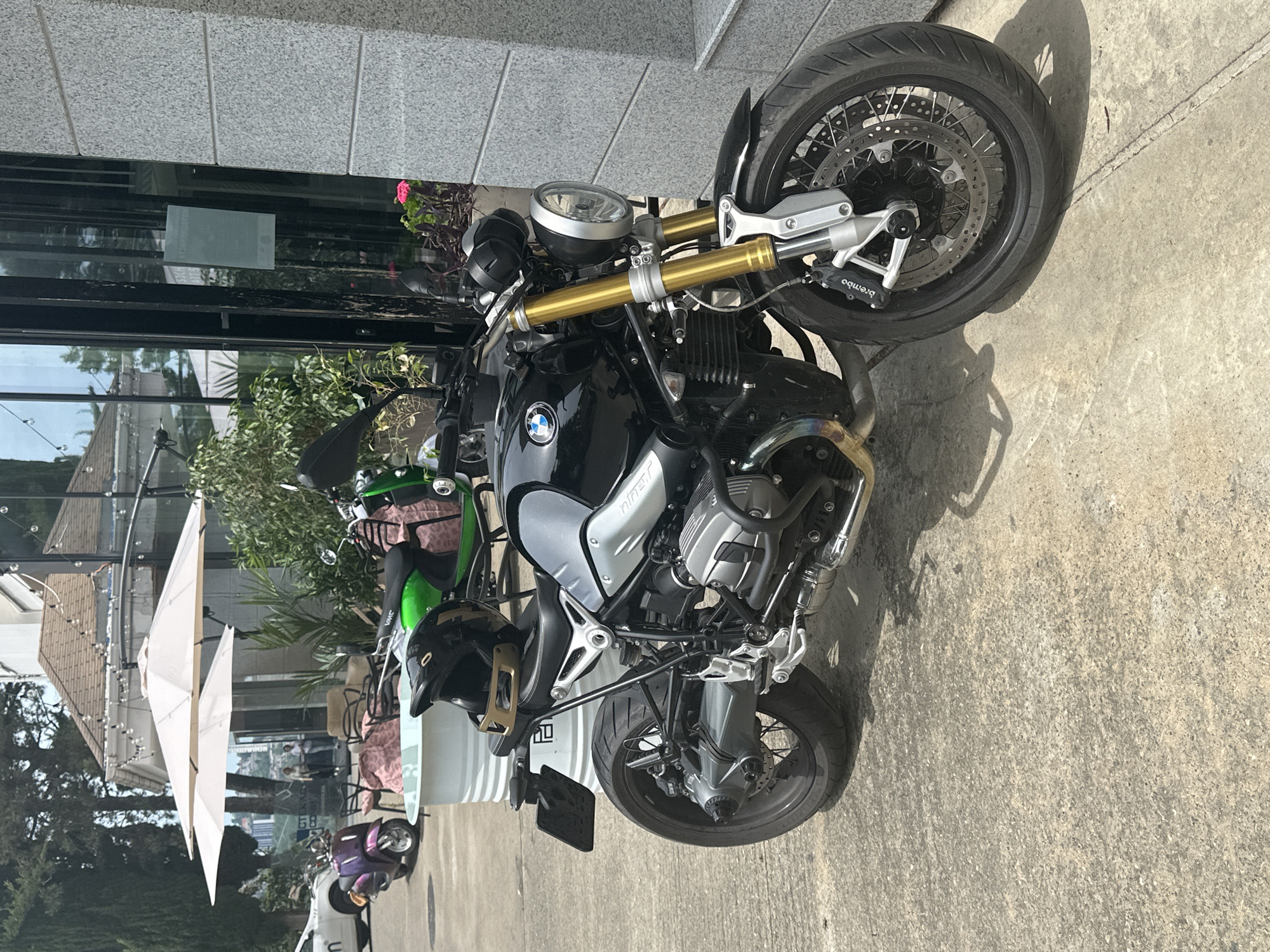 A black BMW motorcycle with silver accents and gold front suspension is parked on a concrete surface. A black helmet with a gold visor rests on the motorcycle's seat. In the background, two purple and green scooters are visible. Behind the scooters, there is an outdoor seating area with chairs and tables, partially covered by two cream-colored umbrellas. A white cylindrical structure is also present. The scene is set against a building with large windows and a gray, textured facade. Trees and foliage are visible in the distance, suggesting an outdoor urban environment.