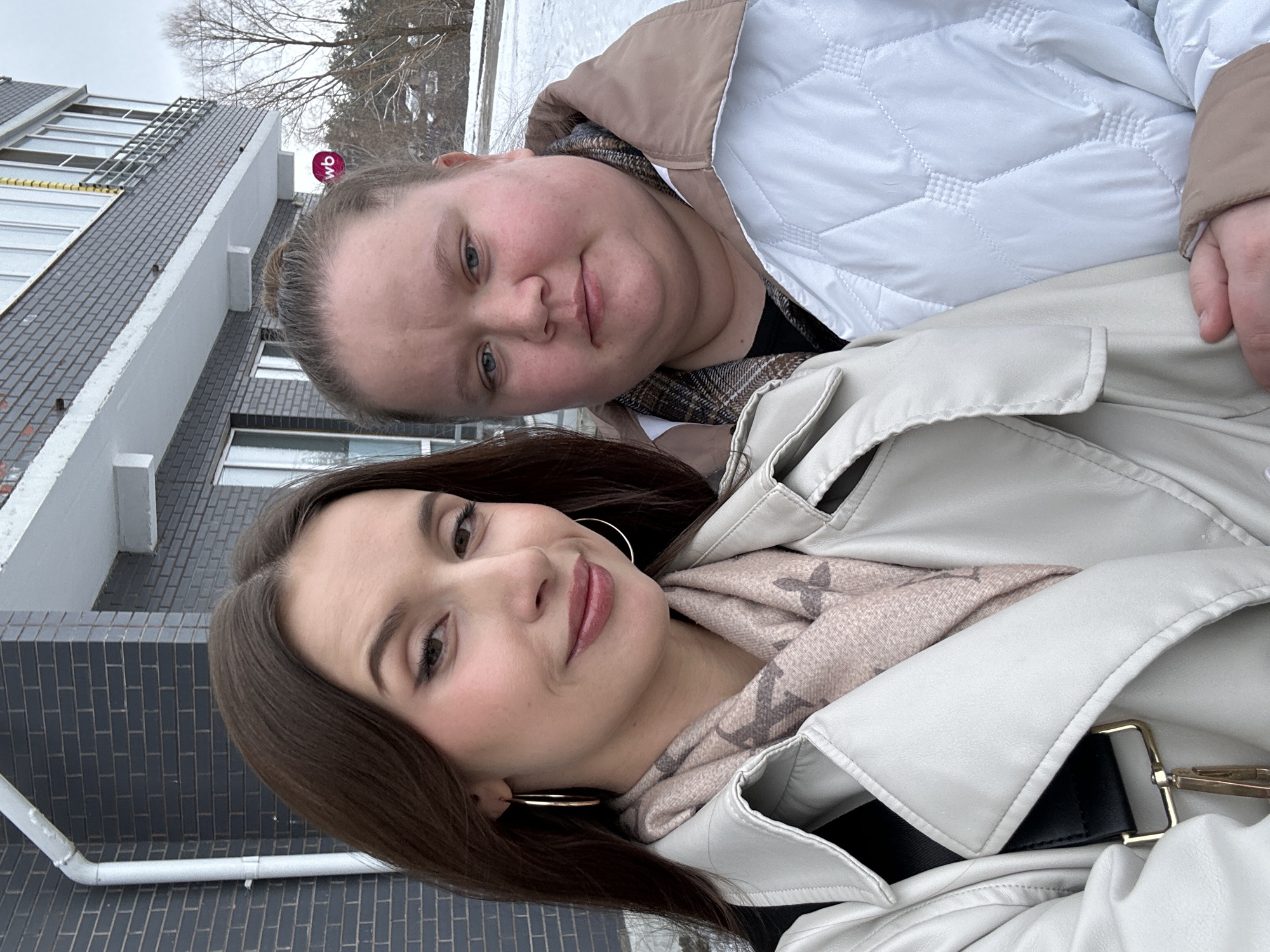 Two women are posing for a selfie outdoors. The woman in the foreground has brown hair, hoop earrings, and wears a light-colored leather jacket over a scarf. The woman behind her has blonde hair and wears a white quilted jacket with a tan hood.
The background includes a building with gray brick and white trim, partially covered in snow. The image was likely taken in the city of Chaikovsky, Russia.