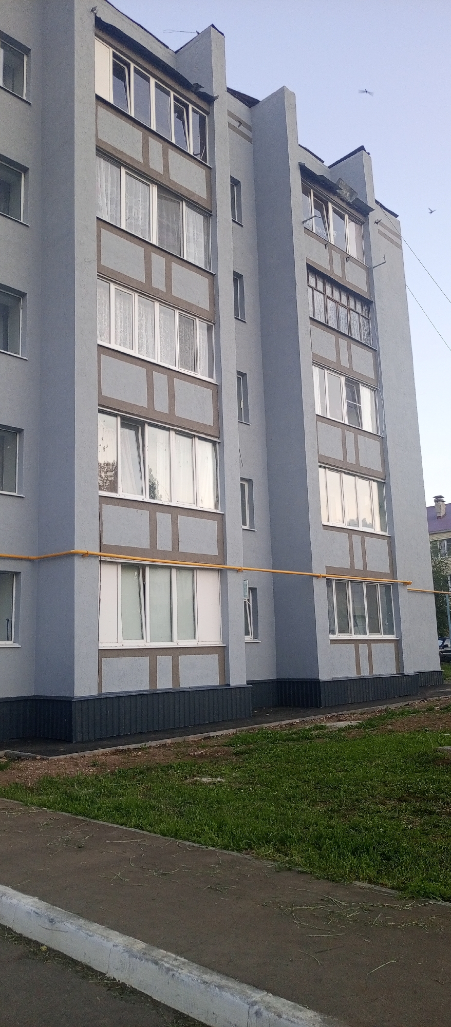 The image shows a close-up view of a modern, multi-story residential building, likely an apartment block, in the city of Agryz, Russia. The building has a light gray facade with dark gray trim at the base and decorative, light brown horizontal and vertical bands on each floor, creating a grid-like pattern. The windows are large and white-framed, with some featuring sheer white curtains. One balcony area on the right side of the building has a darker brown frame and appears to be enclosed with multiple panes of glass. A bright yellow pipe runs horizontally across the middle section of the building's facade. In front of the building is a strip of green grass, and beyond that, a paved walkway with a concrete curb. The sky is clear and a pale blue, with two distant birds visible. The overall impression is of a well-maintained urban residential area.