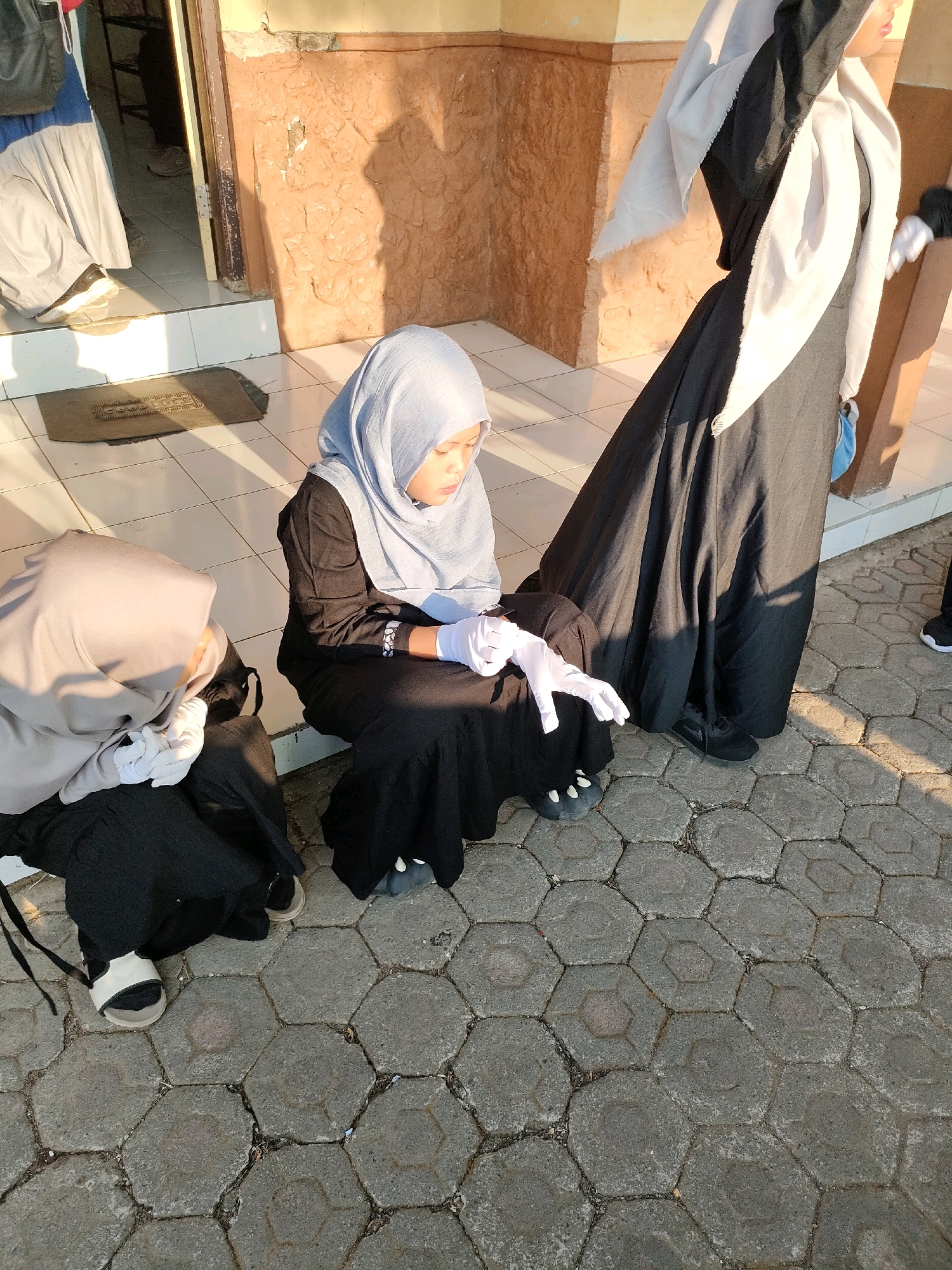 Two individuals wearing hijabs and black dresses are seated on a tiled walkway outdoors. The individual on the left wears a light grey hijab and black pants. The individual on the right wears a light blue hijab, a black dress, and white gloves. A third person, largely obscured by a black abaya and light grey hijab, stands behind and to the right of the seated individuals. The background consists of a textured wall and a doorway. The scene appears to be in Bojong, Indonesia.