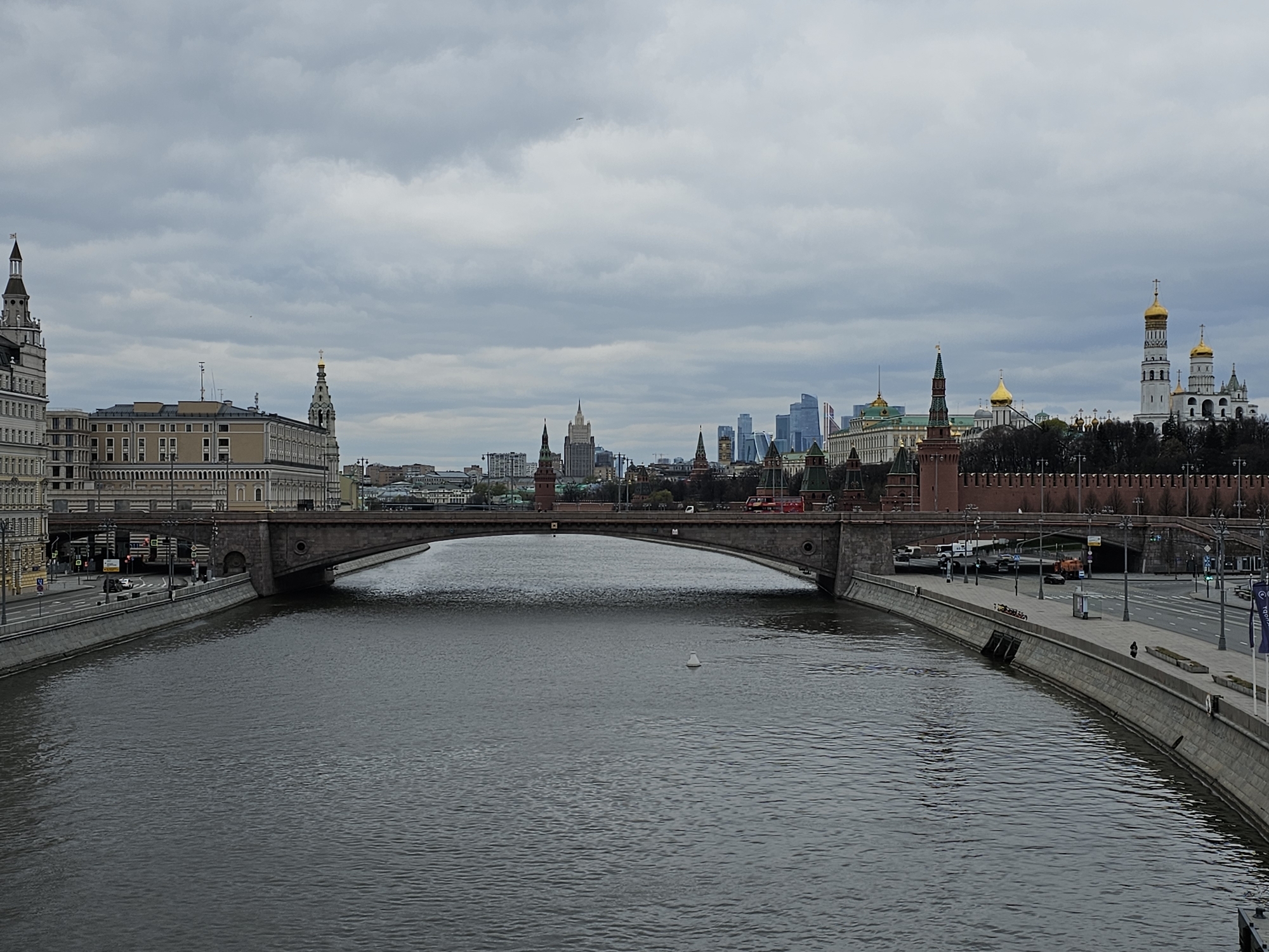 This is a wide-angle shot of a section of the Moskva River in Moscow, Russia. A concrete arch bridge spans the river in the middle ground of the image. On the left riverbank, there is a multi-story building with architectural details. In the background, the towers of the Moscow Kremlin can be seen along with modern skyscrapers. A tall brick wall runs along the right bank of the river. The sky is overcast with dense cloud cover. The river water is calm and gray. There is pedestrian activity on the riverbanks.