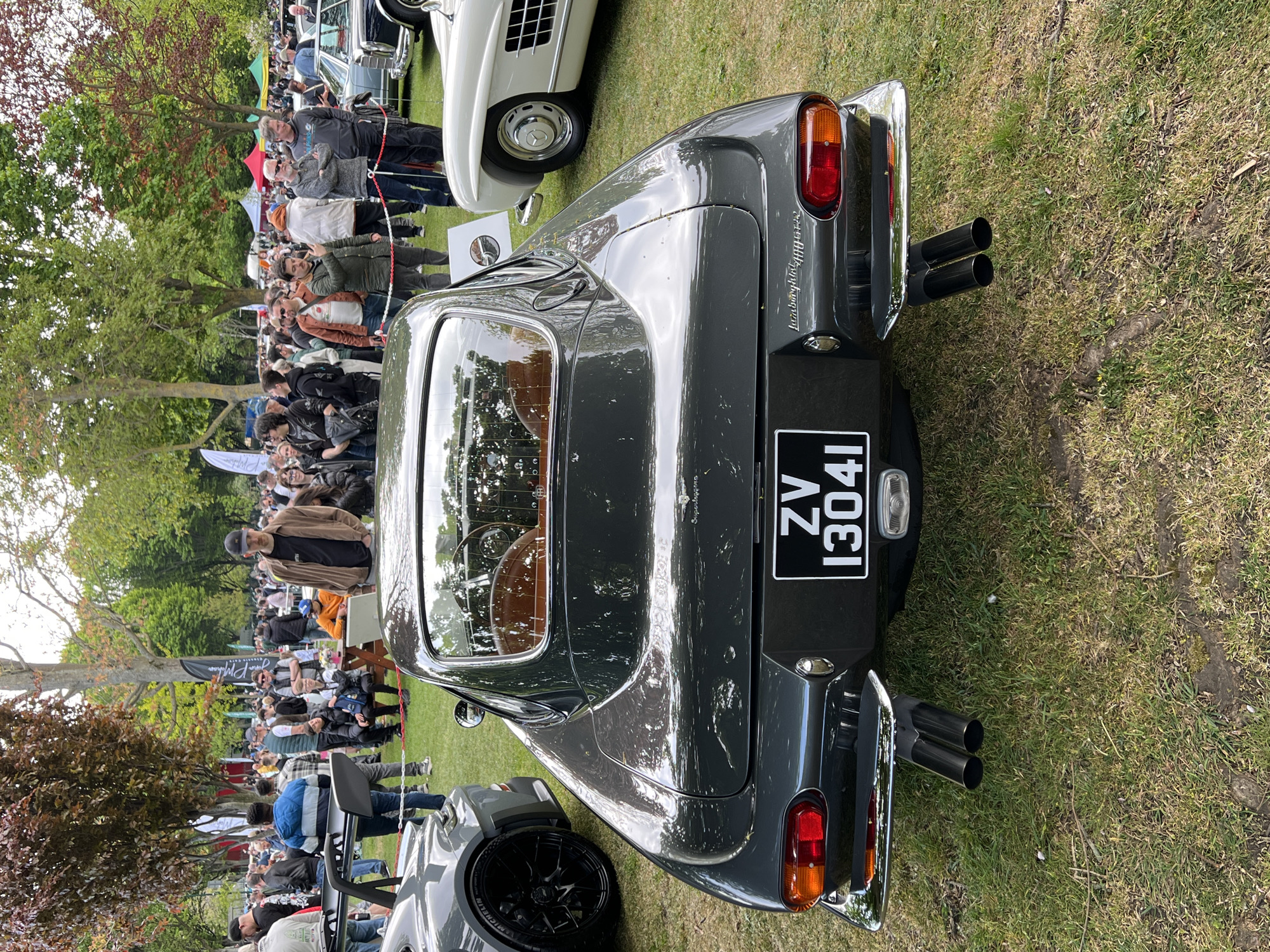 The image depicts the rear of a dark gray Lamborghini 400GT parked on a grass field. The car has chrome dual exhaust pipes on each side and a black license plate with "ZV 13041" in white lettering. The interior of the car is visible through the rear window, showing a brown-colored interior. In the background, there is a crowd of people, a few cars, and some trees. The scene appears to be an outdoor car show or event. The location is not specified.
