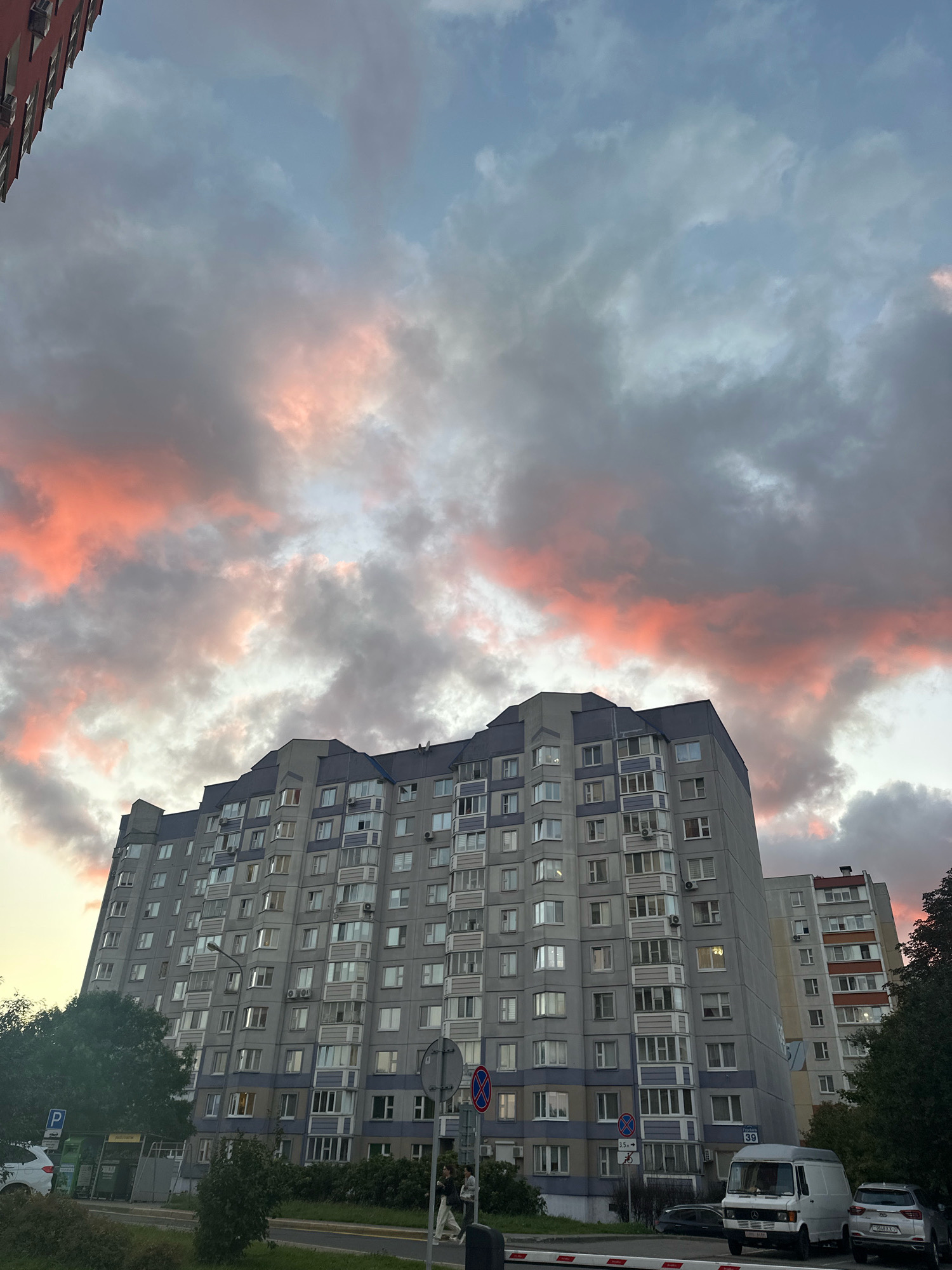 The image shows a multi-story residential building in Minsk, Belarus, under a dramatic sky with pink and gray clouds. The building is predominantly gray with blue accents and numerous windows, some of which are illuminated. In the foreground, there is a street with parked vehicles, including a white van and a silver car. Trees and greenery are visible along the sidewalk. Traffic signs and a parking meter are also present. A few pedestrians are walking on the sidewalk. The sky suggests it might be dusk or dawn.
