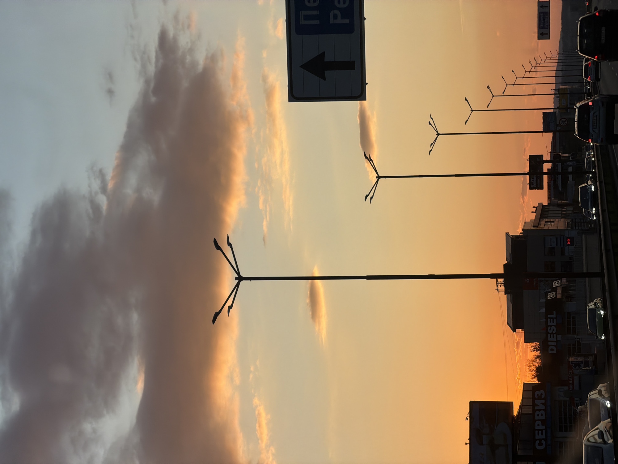 This image captures a wide view of a multi-lane road or highway in Sofia, Bulgaria, during either sunset or sunrise. The sky is a prominent feature, transitioning from a soft blue-grey at the top left to a vibrant gradient of orange, yellow, and pink hues towards the horizon. Scattered clouds, some illuminated by the warm light and others appearing darker and more voluminous, drift across the sky.
On the right side of the image, a continuous line of vehicles, primarily cars and SUVs, can be seen in traffic, suggesting a busy period such as a commute. The cars appear mostly as dark silhouettes against the bright sky, though some show illuminated headlights and taillights. A series of modern streetlights with multiple arms extends along the right side of the road, receding into the distance. These tall structures are silhouetted against the colorful sky.
Alongside the road, various commercial buildings and billboards are visible. These structures are largely in shadow but display visible text. A large overhead traffic sign features a black arrow pointing left and partially visible Cyrillic text, including "Пе" and "Ре" (likely part of a larger destination or road name). On the buildings and billboards, Cyrillic words like "СЕРВИЗ" (SERVIZ - service/repair) are clearly seen, alongside Latin script such as "DIESEL", "AMBULANCE", and "ELITE". A digital clock or display on one building shows "20:09". Further in the distance, another smaller sign with Cyrillic text and an arrow is visible. The scene conveys the bustling atmosphere of an urban highway as day transitions to night or vice versa, with the striking natural light providing a dramatic backdrop to the everyday activity.