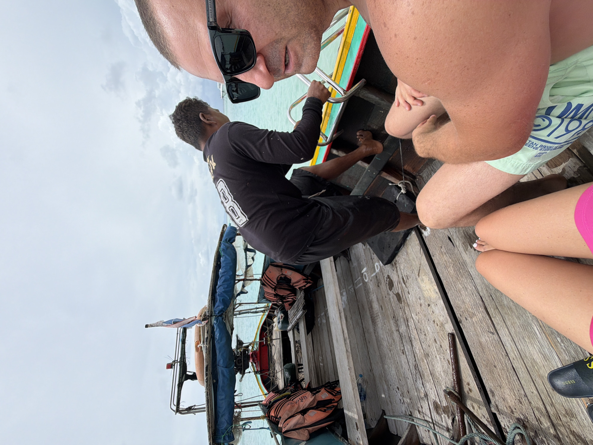 The image depicts a scene on a wooden boat, likely in Ao Nang, Thailand, given the context. In the foreground, a person wearing sunglasses is looking towards the camera, with their right arm resting on their leg. Beside them, a person's legs are visible, one wearing pink shorts and the other a light green patterned swimsuit. In the background, a boatman in a black shirt and shorts is tending to the boat. Life vests are visible on either side of the boat. The boat appears to be on the water, with a cloudy sky overhead.