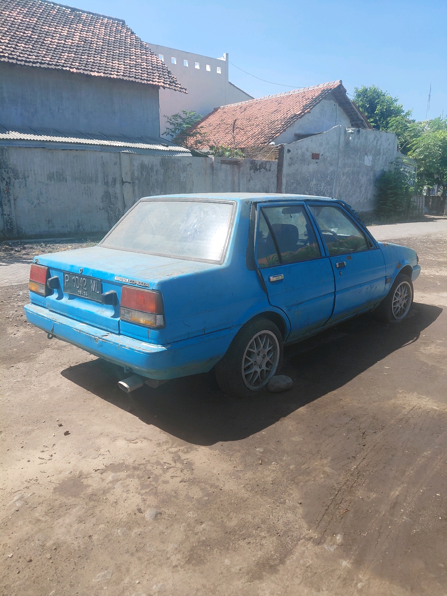 The image shows a blue sedan, a Toyota Corolla, parked on a dirt road in Paiton, Indonesia. The car is old and shows signs of wear, with faded paint and some rust. It has a license plate that reads "P 1042 ML". In the background, there are several buildings with tiled roofs and concrete walls, typical of residential areas in Indonesia. Trees and vegetation are also visible, suggesting a sunny day. The overall scene depicts a quiet, possibly rural, setting in Paiton.