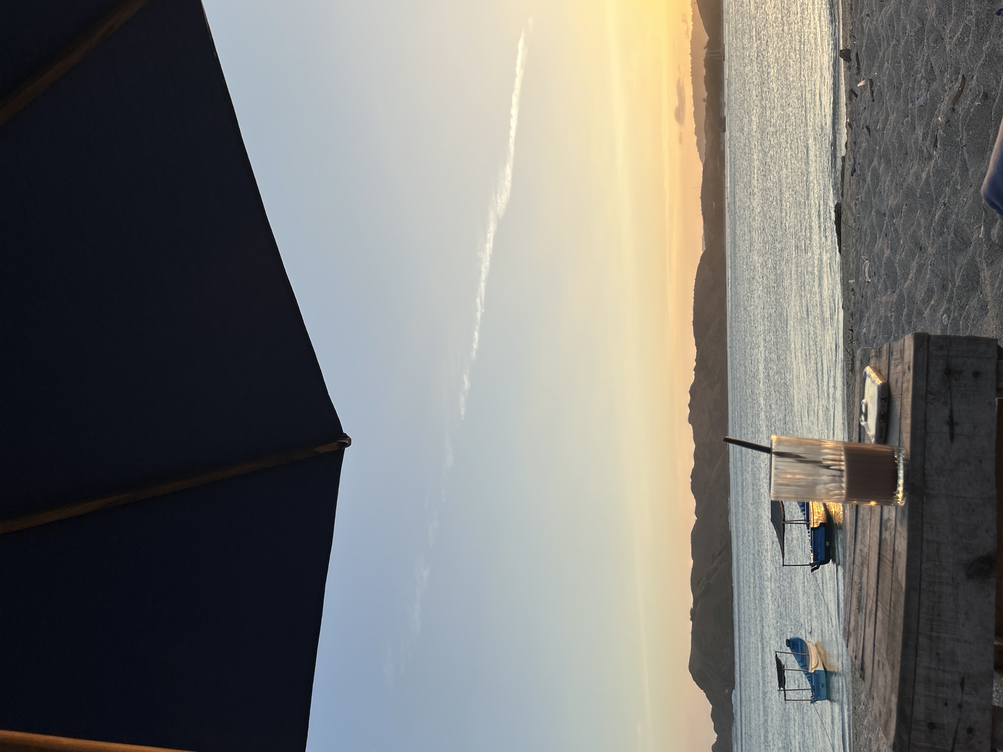 An overhead view of a table with a glass of chocolate drink and a phone on it. In the background, the ocean is visible with several boats and hills. The sky is a gradient of blue and orange, indicating sunset or sunrise. This scene is likely in Kuta, Bali, Indonesia, a popular beach resort town known for its vibrant sunsets and coastal activities.