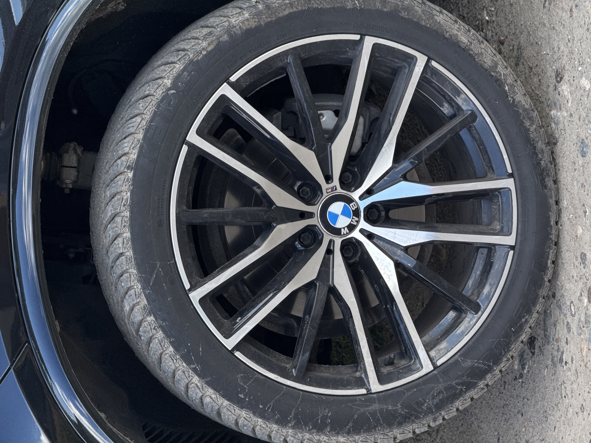 The image is a close-up, eye-level, full shot of a black BMW car wheel, with the car body visible above it. The wheel is a multi-spoke alloy rim with black and silver accents. The tire is black and appears to be moderately worn and dirty, with some dust and debris on the sidewall and tread. The BMW logo is visible in the center of the rim. The wheel is mounted on the front passenger side of the car, as indicated by the positioning of the suspension components visible behind the wheel. The tire and rim are resting on a dirt and gravel surface, suggesting the car is parked outdoors. The lighting suggests it is daytime with some direct sunlight. No people or any other vehicles are visible. The image provides no information about the city of Minusinsk, Russia, or any specific location within it.