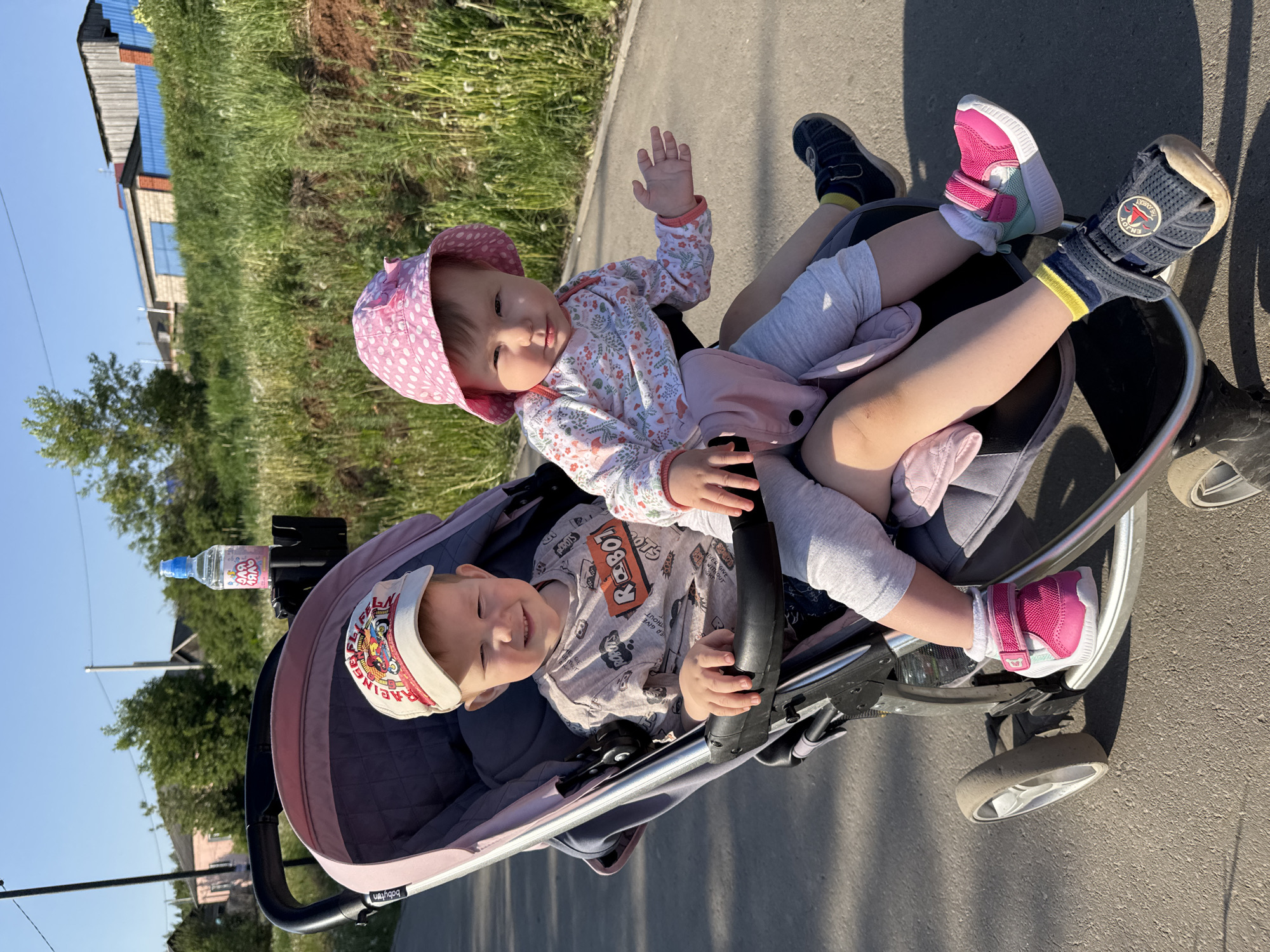 Two young children are riding in a stroller on a paved path in Zav'yalovo, Russia. The child in the stroller is wearing a cap and a patterned shirt, and is smiling. The child on top is wearing a pink polka-dot hat and a floral top, and is waving. Both children have on socks and shoes. A water bottle is attached to the stroller. The background shows greenery and buildings.