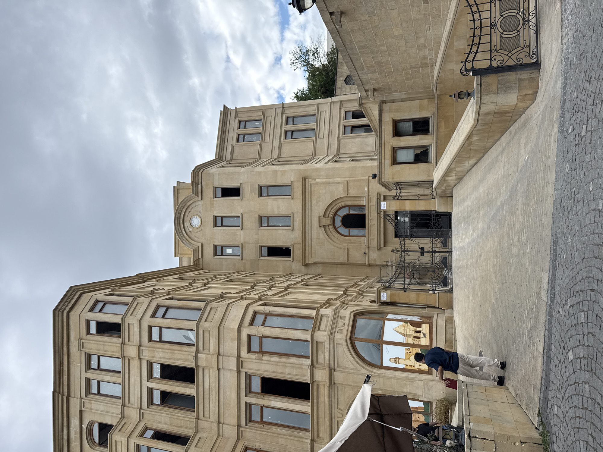 This media file shows a street view of a building in Sabail Raion, Azerbaijan. The building is made of light-colored stone with multiple floors and many windows. The architecture is ornate with decorative trim and arched windows. The sky is overcast with clouds, suggesting a potentially cool or damp day.
In the foreground, a person is standing on a paved walkway, leaning slightly forward. They are wearing a dark blue shirt and light-colored pants. To the left and partially in the foreground, a white umbrella or canopy is visible, suggesting a possible market or outdoor seating area. The ground is paved with cobblestones and large stone tiles.
The building itself features several distinct architectural elements, including a large arched window on a curved section of the building that reflects other structures, possibly with minarets. There is also a clock visible on one of the upper sections of the building. Decorative metalwork, possibly a gate or railing, is present on an upper level.
The overall impression is that of a historical or well-maintained urban environment. The lighting is diffused due to the cloudy sky. There is no visible text in the media file. The scene appears to be a quiet moment, with the person in the foreground being the only clearly identifiable human subject actively engaged in the scene. The architecture hints at a location with a rich history and distinct cultural influences.