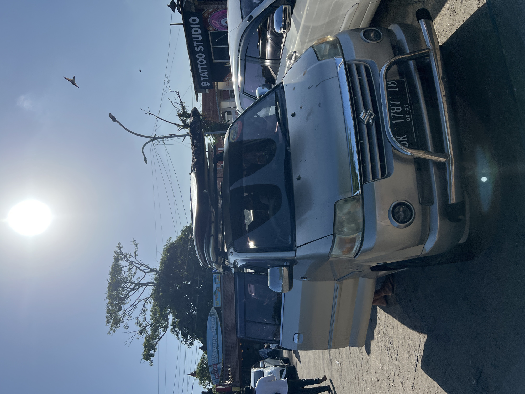 A silver van with surfboards on the roof rack is parked on a paved surface in front of a "Warung Mody" restaurant. A person is standing with the van door open, partially visible behind the door. To the right, a white car is parked next to the van. In the background, a "Tattoo Studio" sign is visible on a building. The scene is outdoors on a sunny day, with power lines and trees in the background. A bird is flying in the bright sky. The license plate on the van reads "DK 1787 IU".