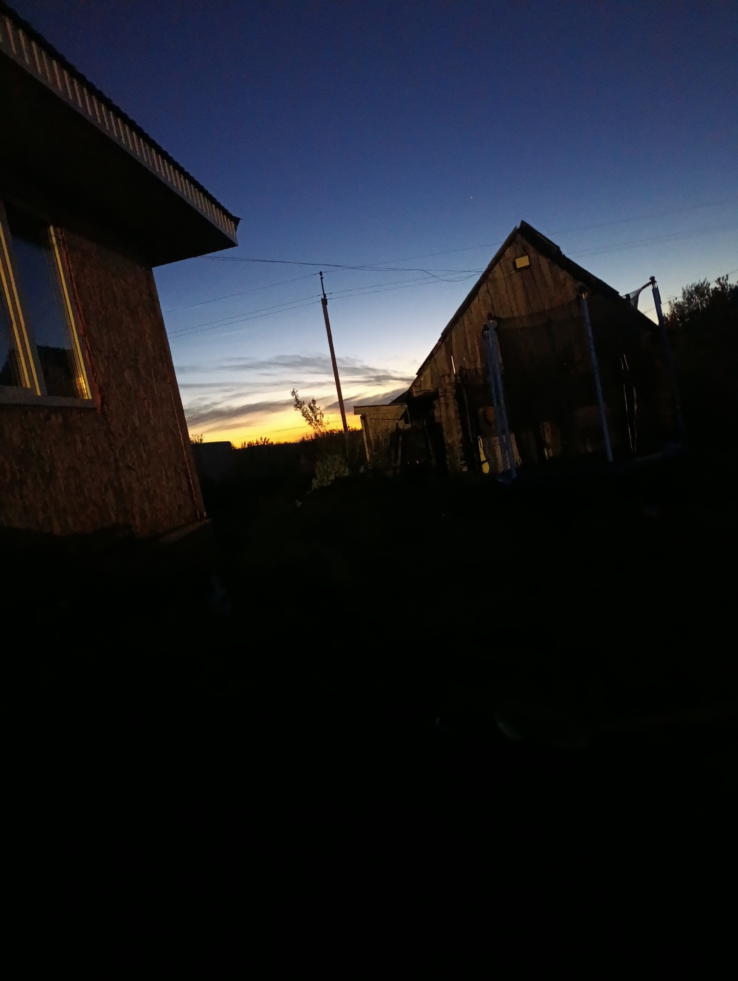 The image shows an outdoor scene at dusk, with a deep blue sky transitioning to lighter hues near the horizon. In the foreground on the left, a part of a building with a brown, textured facade and a sloped roof is visible. A window with yellow trim reflects the sky. To the right, a wooden structure, possibly a shed or a small house, stands with a gabled roof. Its dark silhouette is contrasted against the fading light. Power lines and a utility pole are also present in the background. The ground is dark and appears to be covered in vegetation. The overall setting suggests a rural or semi-rural environment. The location is identified as Buzov'yazy, Russia.