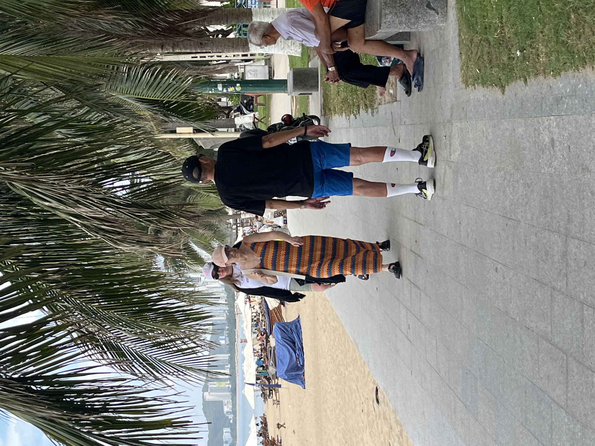 The image depicts a beachfront promenade in Nha Trang, Vietnam, characterized by a sandy beach, palm trees, and a tiled walkway. On the right side of the walkway, two men are seated on a stone bench. One man, wearing a light purple collared shirt and dark pants, faces away from the camera, looking towards the beach. The other man, partially visible, wears an orange shirt and dark shorts. In the center of the image, two women walk along the promenade. The woman in front wears a long, orange and striped maxi dress with a white strap over her shoulder and dark sandals. She is followed by another woman wearing a white t-shirt, dark shorts, and a black jacket, with a light-colored baseball cap and sunglasses. On the left side of the walkway, a man wearing a black t-shirt, blue shorts, white socks, and athletic shoes walks away from the camera. Behind him, a blue tarp covers an object on the sand. Further in the background, beach chairs and umbrellas are visible on the sandy beach, with a cityscape and ocean in the distance. The environment suggests a warm, tropical setting.