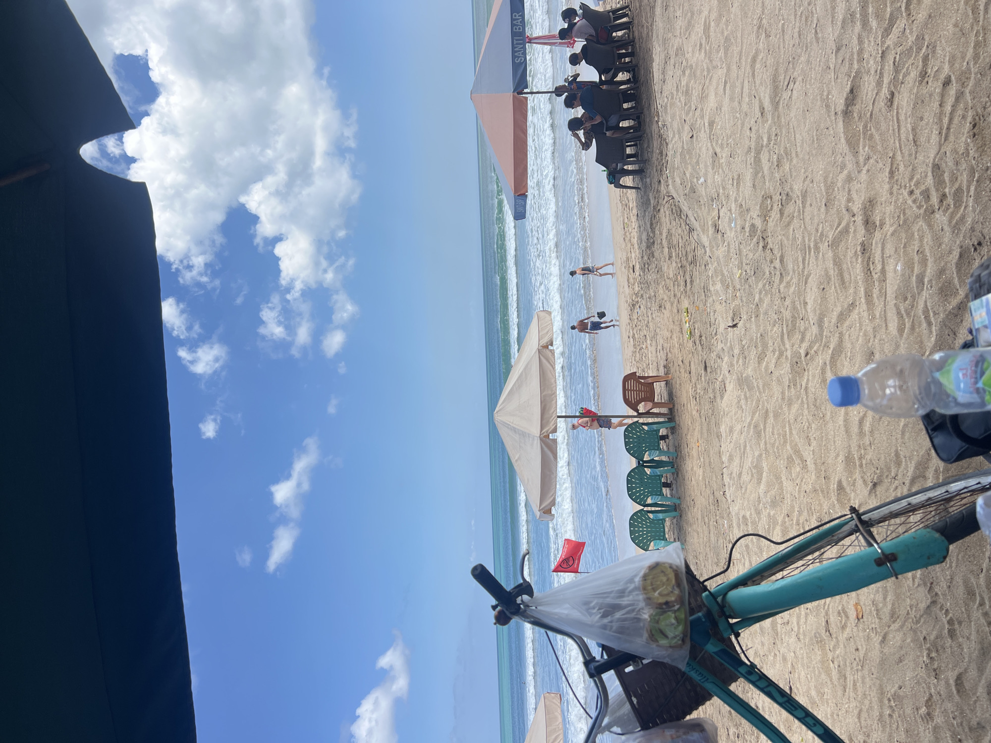 The image depicts a beach scene in Kuta, Indonesia, on a sunny day with scattered clouds. In the foreground, a turquoise bicycle is parked on the sand, with a plastic bag containing food items hanging from its handlebars. A bottle of water rests on a bag next to the bicycle.
Further back, beach chairs and umbrellas are set up on the sand. Several people are seated under one of the umbrellas, which has "SANTI BAR" written on it. In the distance, people are walking on the beach and in the shallow water. The ocean waves are visible, and the horizon line separates the blue sea from the sky. A red flag, possibly indicating a warning or safety notice, is planted on the beach. The overall atmosphere is one of a relaxed beach day with people enjoying the seaside.