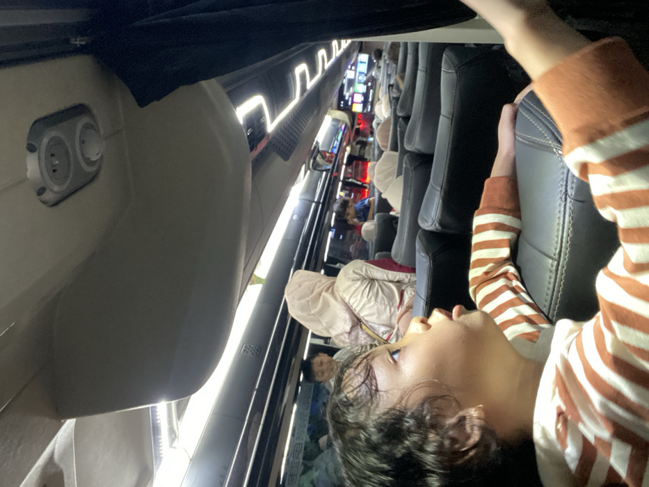 The image depicts the interior of a passenger bus, reportedly located in Pandaan, Indonesia. The view is from a seat on the right side of the bus, looking forward. In the immediate foreground on the left, a young child with dark, curly hair is seated, wearing a white and brown-striped long-sleeved shirt. The child's head is turned to the right, mouth slightly open, looking upwards and forward. A small gold earring is visible in the child's left ear. The child's left arm rests on the armrest of a dark, upholstered bus seat.
In the midground, directly behind the foreground child, a young boy in a light-colored shirt is visible, looking towards the front. To the right of this boy, an adult female is seated facing away, wearing a light-colored hijab and attire, with a shoulder bag strap visible. Further into the midground and background, multiple other passengers are seated in dark, high-backed seats, some wearing head coverings. A young child in a blue shirt is partially visible on the right side of the cabin, further back.
The bus interior features overhead luggage compartments, linear LED lighting strips running along the ceiling, and individual overhead service panels containing what appear to be power outlets or light controls. A black curtain is partially visible on the right side of the frame. Towards the front of the bus, a monitor displaying colorful graphics is visible. The scene is illuminated by the bus's interior artificial lighting.