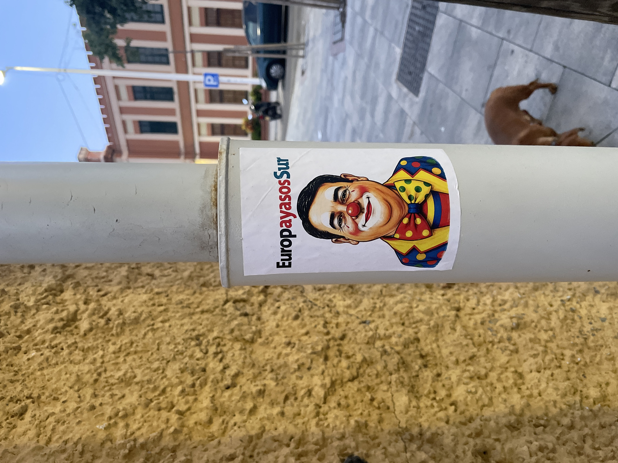 The image shows a sticker on a lamppost in La Línea De La Concepción, Spain. The sticker features a cartoon drawing of a man with clown makeup, including a red nose and a colorful outfit with a large bowtie. The text "EuropayasosSur" is printed above the man's head. The background of the image shows a textured yellow wall, a sidewalk, a building with red and white stripes, a parking sign, a scooter, a car, and a dog walking on the sidewalk. The sky is a light blue, suggesting it is either early morning or late afternoon.