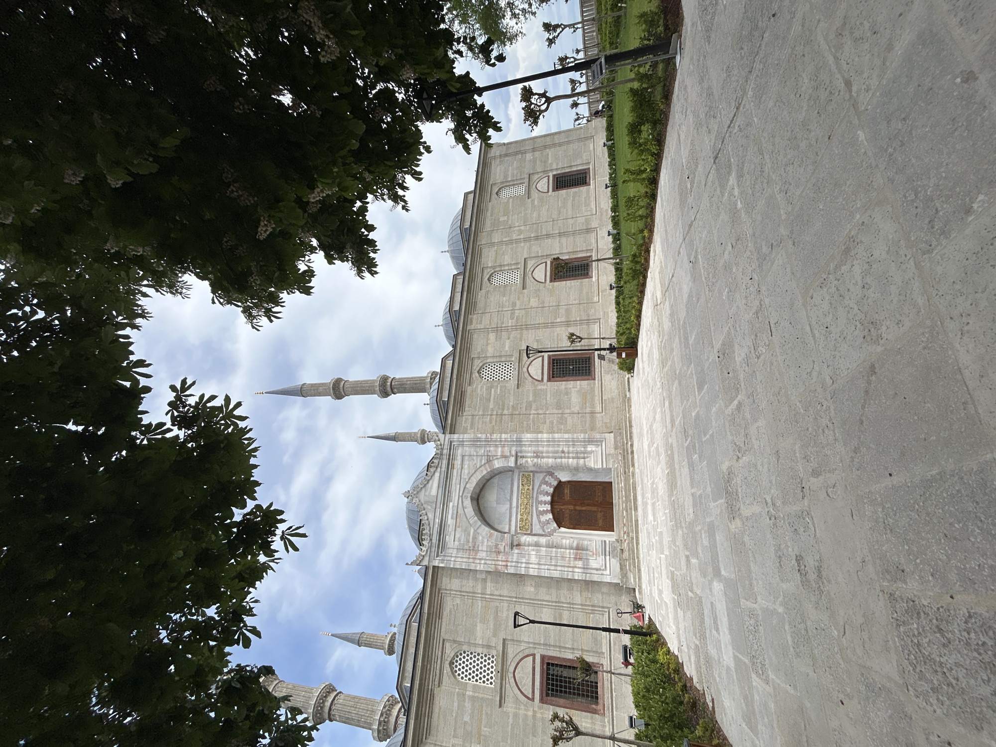 The image is of the Selimiye Mosque in Edirne Merkez, Turkey, shot from a low angle, and rotated 90 degrees counter-clockwise. Part of a cobblestone pathway is in the foreground on the right. The mosque's architecture includes multiple minarets, domes, arched doorways, and latticed windows. Tree foliage obscures the upper-left corner of the frame, with partial views of other trees in the background. Utility poles and green shrubbery are visible between the pathway and mosque. The sky is cloudy.