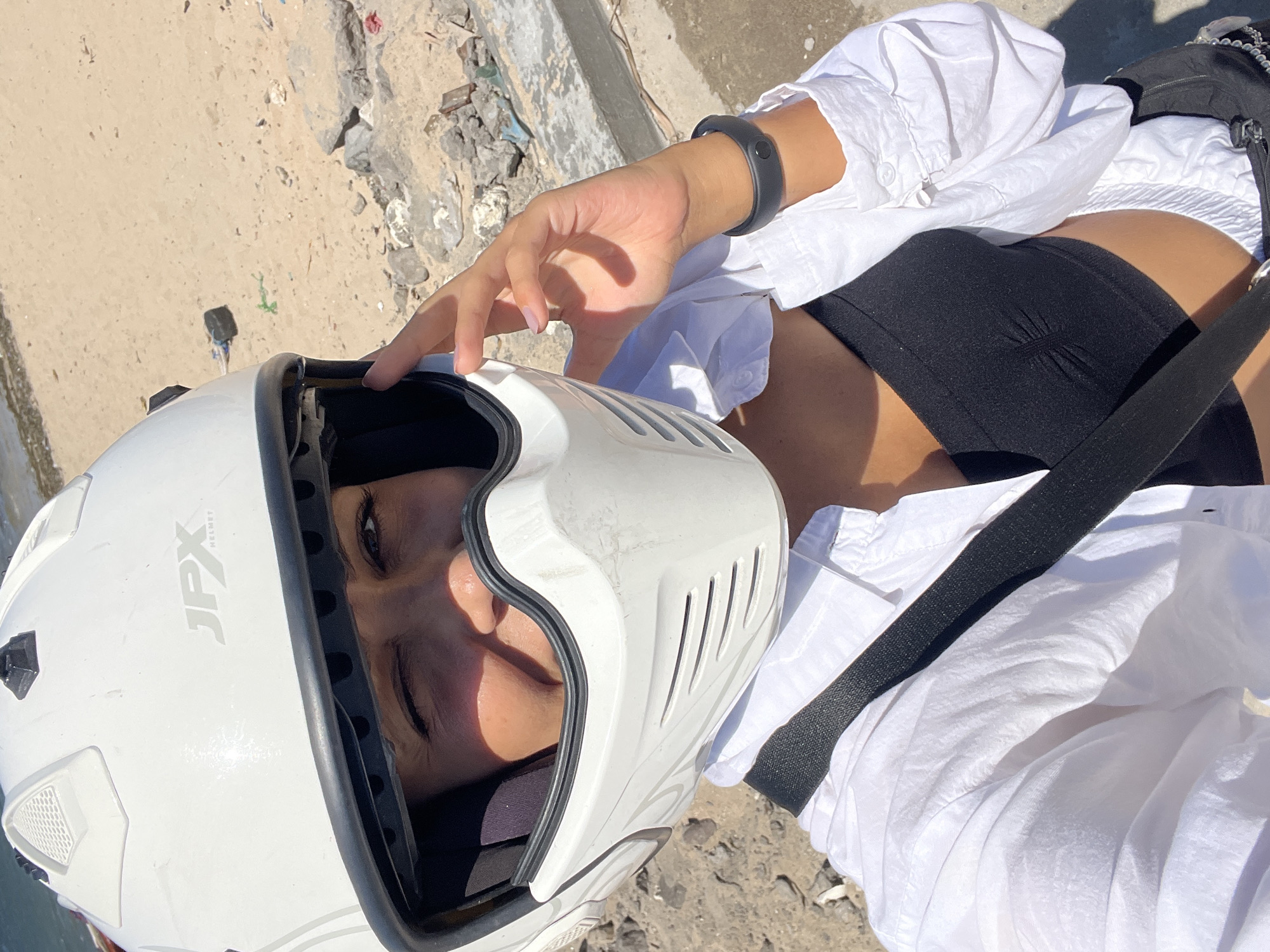 A person wearing a white full-face helmet with the brand name "JPX" visible is pictured in a close-up, high-angle shot. The helmet's visor is partially open, revealing the person's face inside. The person is wearing a black tank top and a white button-down shirt. A black wristband is on their left wrist. A black strap crosses their body. The background shows a tan, sandy surface and a concrete ledge with debris. The location is in the city of Unknown, Indonesia.
