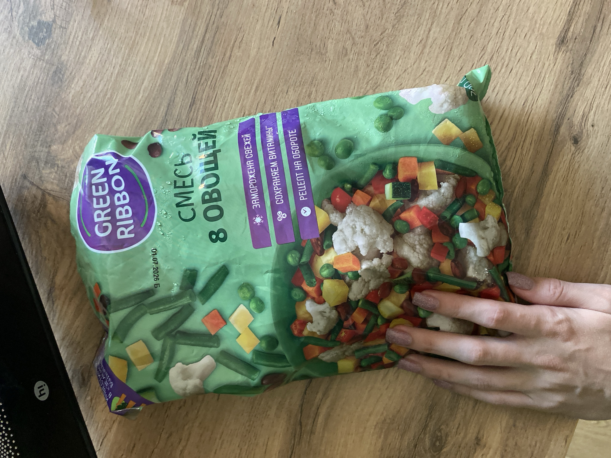 A hand with pink sparkly nail polish rests on a package of frozen vegetables, labeled "GREEN RIBBON", on a wooden surface. The package is predominantly green with colorful illustrations of vegetables and text in Russian, including "СМЕСЬ 8 ОВОЩЕЙ" (Mix of 8 vegetables). Three purple text boxes on the right side of the package contain text that reads "ЗАМОРОЖЕНА СВЕЖЕЙ" (Frozen Fresh), "СОХРАНЯЕМ ВИТАМИНЫ" (We Keep Vitamins), and "РЕЦЕПТ НА ОБОРОТЕ" (Recipe on the back). The background is a wooden table with a dark, glossy object in the upper left corner that displays the letters "hi". The image appears to be taken indoors.