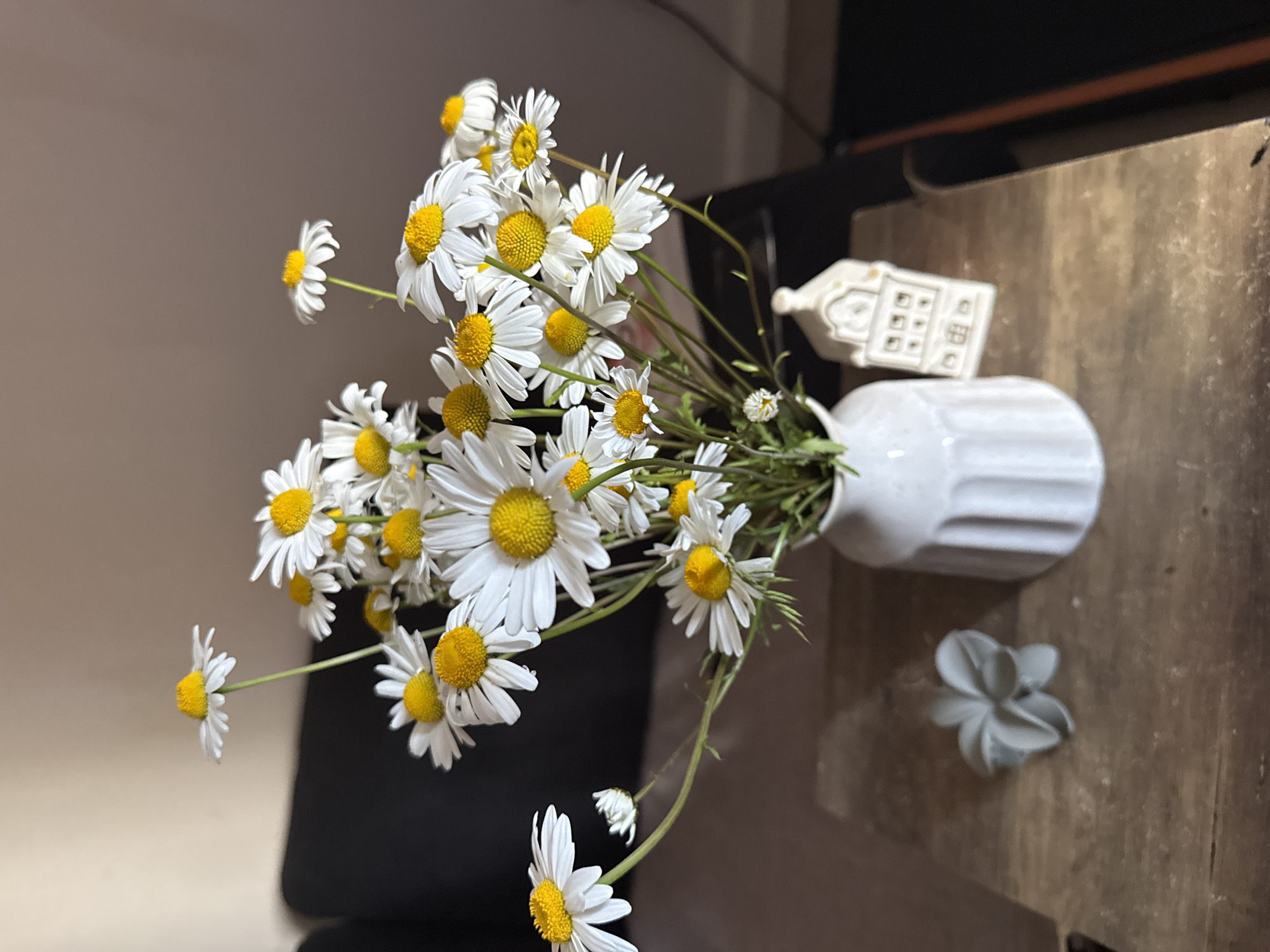 A bouquet of daisies in a white vase sits on a wooden surface next to a small decorative house and a flower-shaped object. The backdrop features a blurred wall and a dark object, possibly a piece of furniture. The scene appears to be indoors. There is no indication that this scene is located in Great Yarmouth, United Kingdom. There are no people visible in the image.