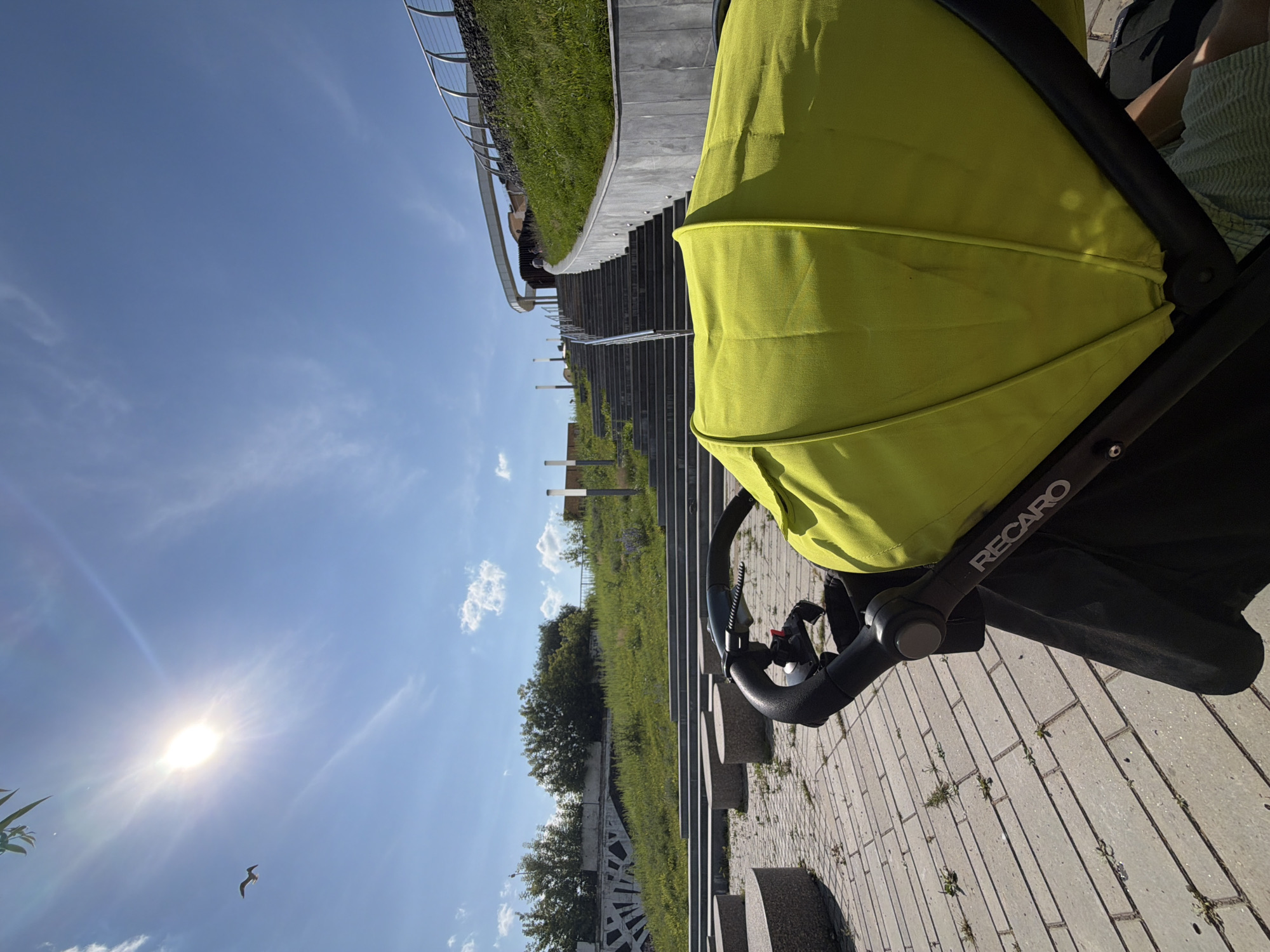 An outdoor scene depicts a bright, sunny day with a clear blue sky and some wispy clouds. In the foreground, the handle and bright yellow canopy of a stroller are visible, with the brand name "RECARO" clearly printed on the handle. The stroller is positioned on a paved pathway made of light-colored rectangular stones, with some sparse weeds growing between them.
In the midground, a series of wide, concrete steps leads upwards, flanked by grassy slopes and modern metal railings. Some cylindrical stone seating elements are scattered on the paved area to the left of the steps. Beyond the steps, trees and foliage form a green backdrop. In the far distance, a structure with a curved roof and visible supports is situated on a hill, and a person can be seen walking up the stairs.
The sun is high in the sky, casting strong light and shadows. A bird is captured in flight against the bright sky. The scene appears to be in a park or recreational area, possibly a city promenade or public space with architectural elements designed for pedestrian access and leisure. The location is identified as Votkinsk, Russia.