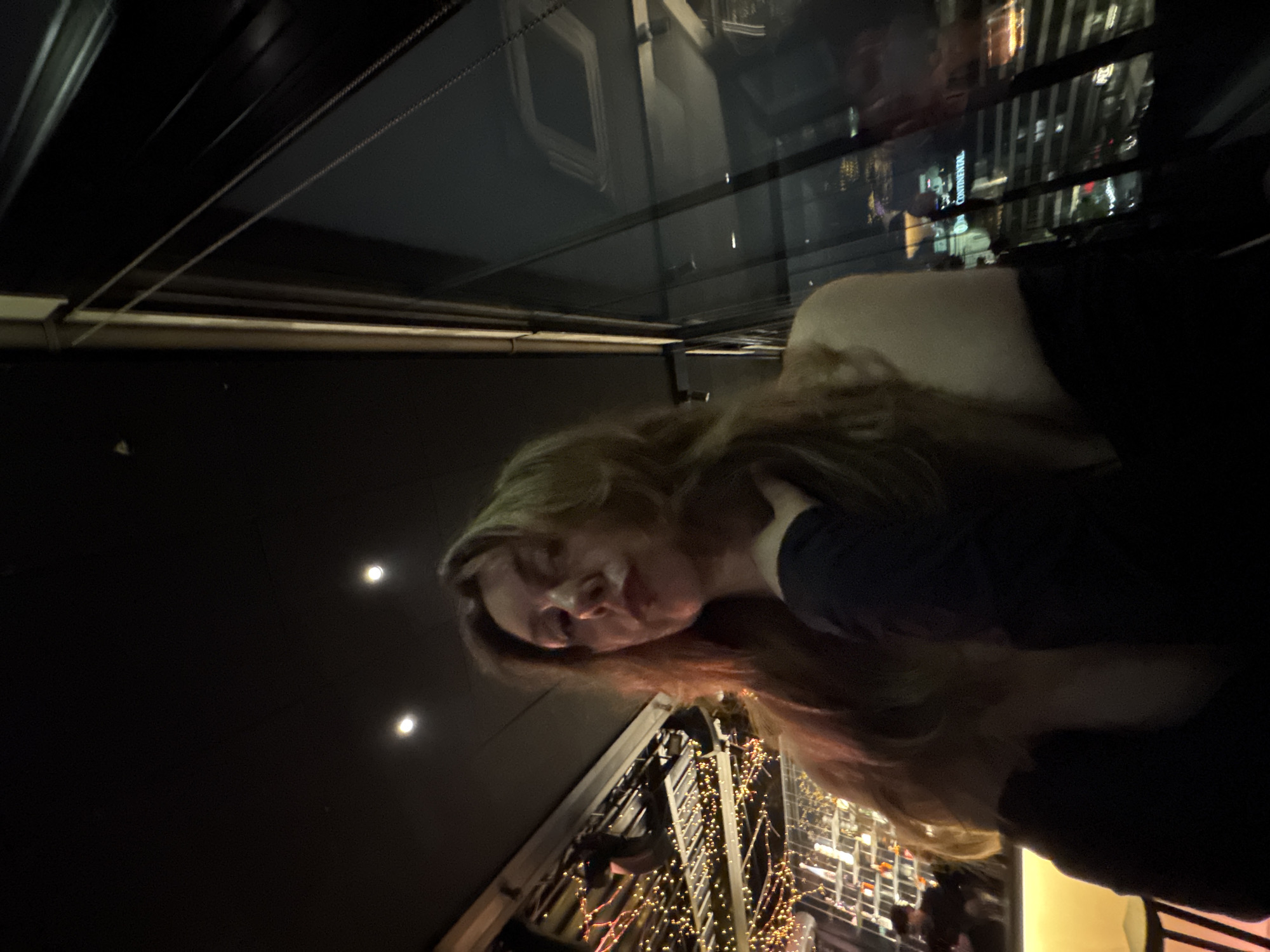 The image shows a woman with long, blonde hair, wearing a dark-colored shirt, standing on a balcony at night. Reflected in the balcony's glass is a cityscape lit up with lights. There is a building decorated with string lights. The exact location within the city is not discernible.