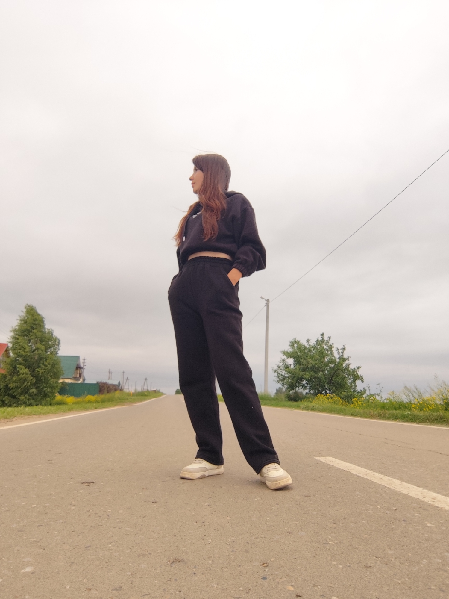 A young woman with long brown hair stands on an asphalt road, looking to her left. She is wearing a black hooded sweatshirt and black sweatpants. The road stretches into the distance, flanked by green grass and trees. In the background, there are houses and telephone poles, suggesting a rural or semi-rural setting. The sky is overcast, with a pale, diffused light. The image appears to have been taken in or near the city of Fokino, Russia.