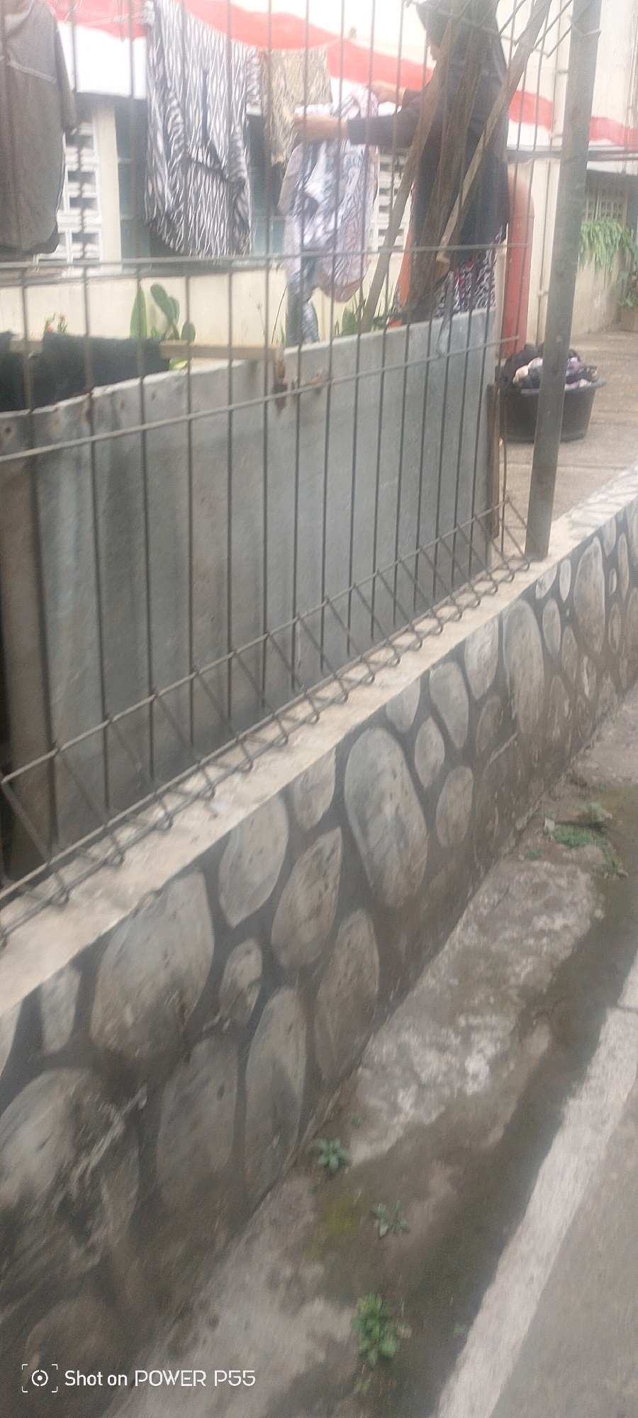 An outdoor scene in a city shows a chain-link fence. Beyond the fence, clothes are hanging on a clothesline, some of them appearing to be patterned. A person is visible behind the clothes, and a black bucket filled with clothes is on the ground to the right. The fence is supported by a concrete wall that is decorated with painted stones. Some patches of green vegetation are visible on the ground to the right of the wall. The scene is dimly lit and appears to be overcast.
