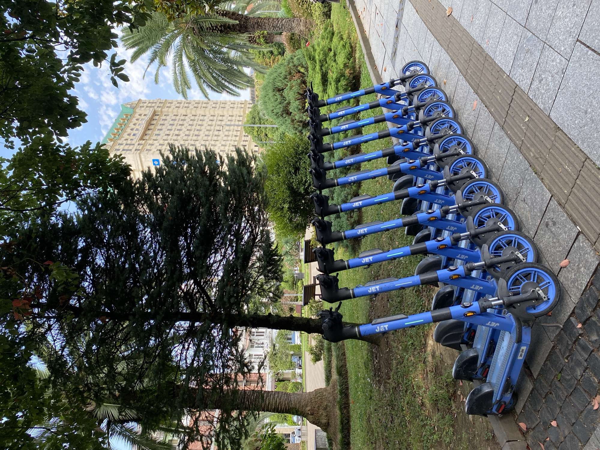 A line of nine blue JET branded electric scooters is parked along a sidewalk in Batumi, Georgia, with a large, cream-colored, multi-story building with a green-tiled roof visible in the background. The scooters are positioned in a grassy area beside the paved walkway. The building, which appears to be a hotel, has many windows and decorative architectural elements. Palm trees and other lush greenery surround the scooters and the building. The sky is partly cloudy.