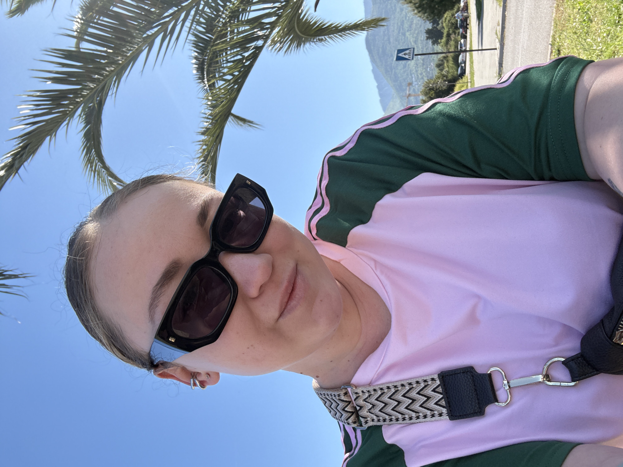 A woman in sunglasses and a pink and green shirt is posing in front of a palm tree. The location is on a road in Bar, Montenegro. A pedestrian crossing sign is visible in the background. The sky is blue. She is wearing a patterned shoulder strap.
