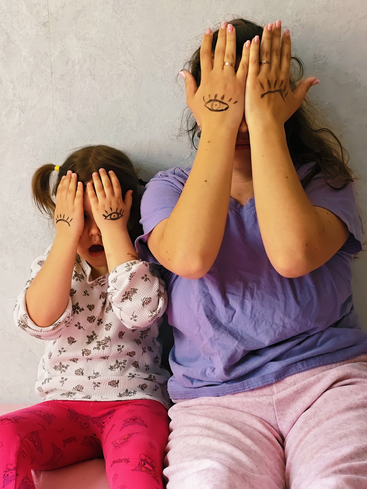 Two females, an adult and a young child, are captured indoors against a light gray, textured wall in Chaikovsky, Russia. Both are covering their faces with their hands, which have playful eye drawings on them, creating a whimsical effect of "eyes" peeking through their fingers.
The adult, positioned on the right, has long dark hair and is wearing a purple short-sleeved top and light pink sweatpants. Her hands, adorned with pink nail polish and rings on some fingers, feature a more detailed eye drawing with a pupil on her left hand (from the viewer's perspective) and an outline with prominent lashes on her right hand.
The child, on the left, has brown hair styled in pigtails with yellow hair ties. She is wearing a white long-sleeved top with a subtle floral pattern and vibrant pink leggings that appear to have cartoon characters on them (though specific characters or text are not clearly discernible). Her hands also bear drawn eyes, with one showing a distinct eye with a pupil and the other featuring prominent eyelashes. Her mouth is open, giving the impression of surprise or engagement in a playful act.
The scene suggests a casual, indoor setting, possibly at home, with a lighthearted and interactive moment between the two individuals. The drawn eyes on their hands add a unique and humorous element to the image.
