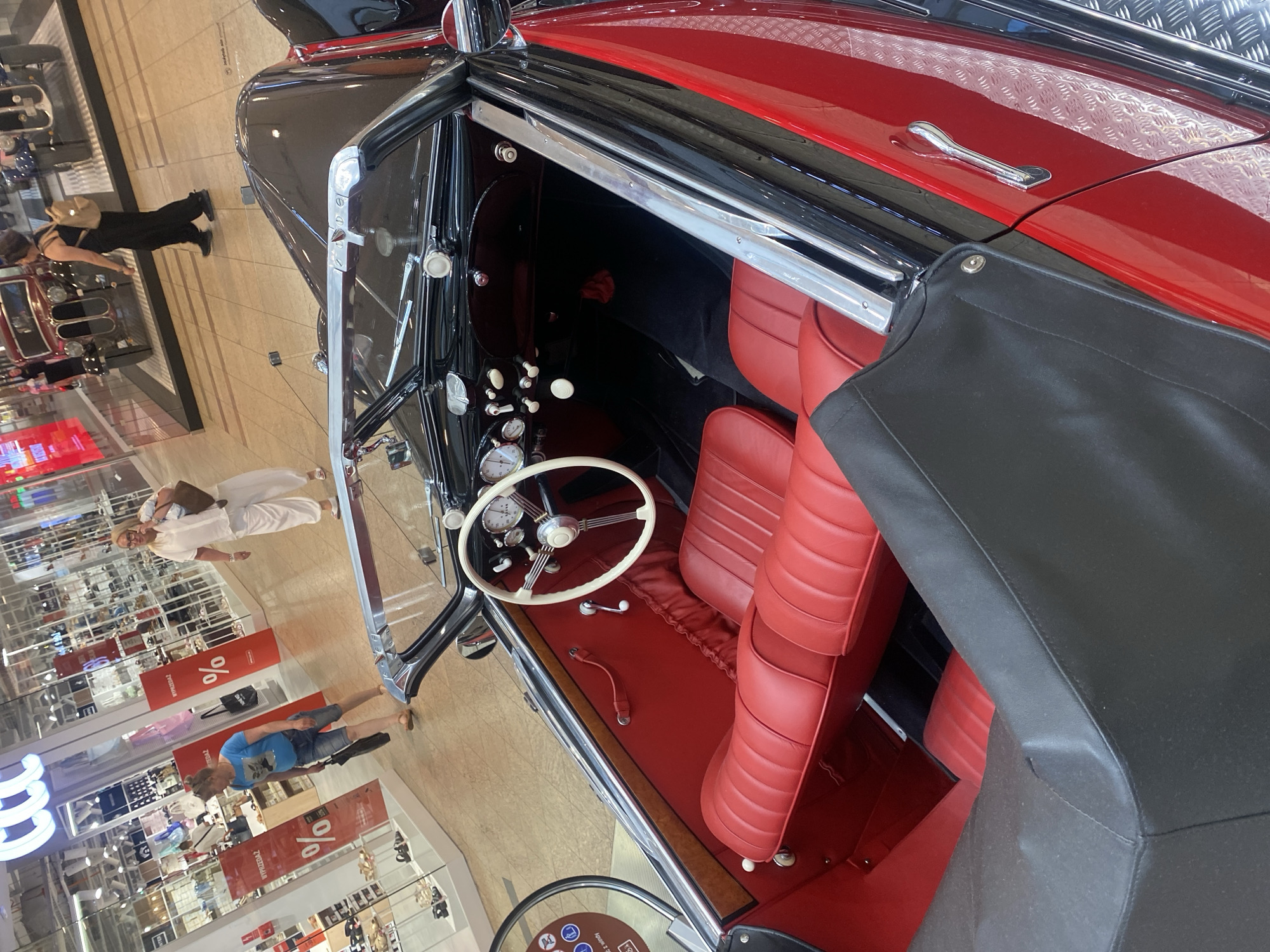 In a retail setting, potentially a mall in Łódź, Poland, a vintage black and red convertible car is on display. The car's interior is a striking red, featuring plush leather seats and a classic white steering wheel. The dashboard is equipped with multiple gauges and white knobs. A black soft-top is partially covering the passenger area.
Several people are present in the background, browsing in shops. One woman in a white outfit stands near the car, while another woman in a blue t-shirt and denim shorts is further away. Red banners with a "%" symbol are visible in the shopping area, suggesting a sale or promotional event. The scene appears to be indoors, well-lit, and possibly during daytime. The overall atmosphere suggests a leisurely shopping or exhibition experience. No specific time of day or weather conditions can be determined from the image.