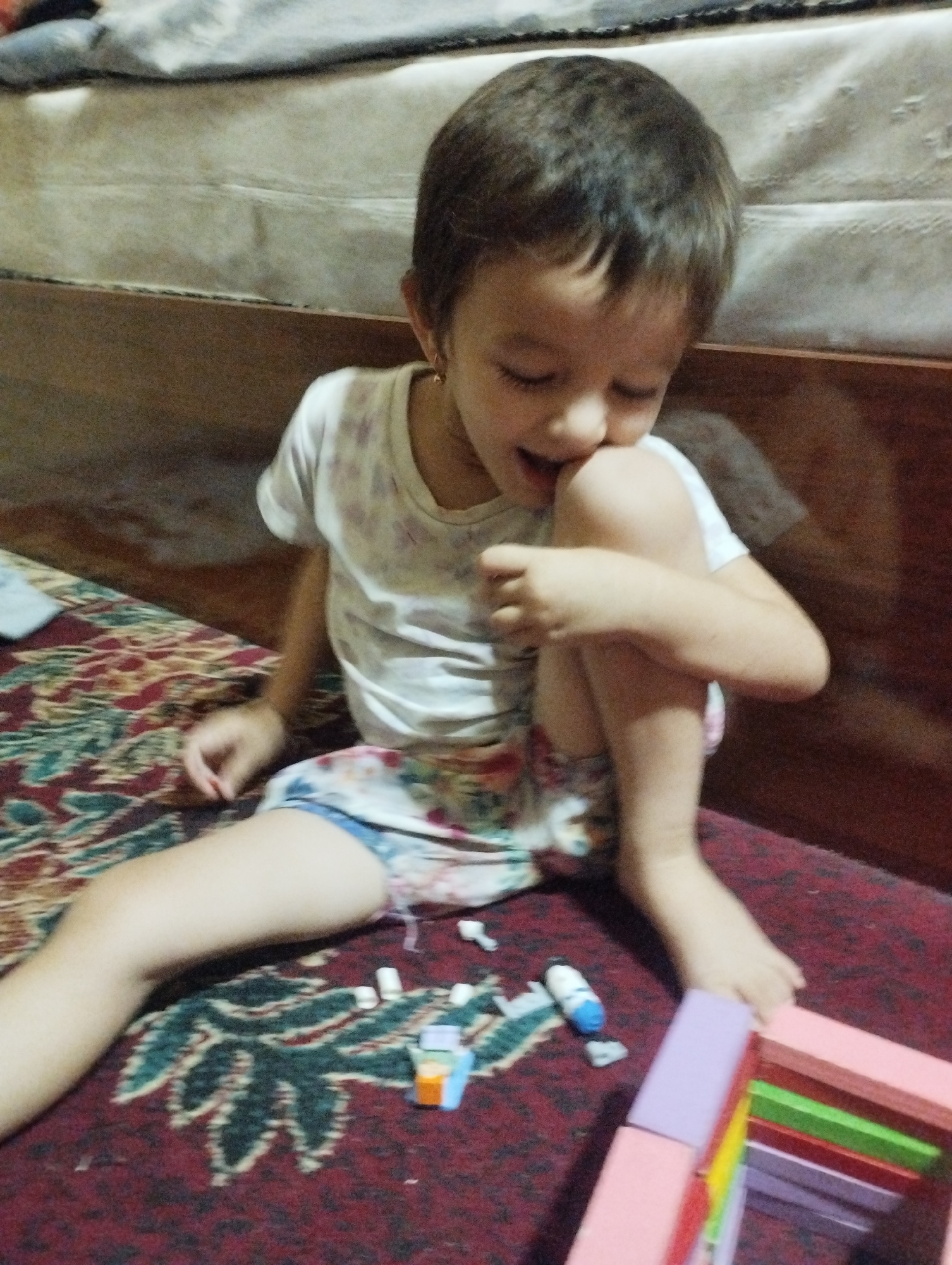 A young child is sitting on a patterned rug, with their knees pulled up to their chest. The child is wearing a white t-shirt with a colorful pattern and patterned shorts. They are looking down and appear to be playing with small objects on the rug, including what looks like building blocks and cylindrical pieces. In the foreground, a pink frame with colorful rods is visible. The background shows a bed with a beige headboard and a wooden structure. The lighting suggests an indoor setting, likely a home.