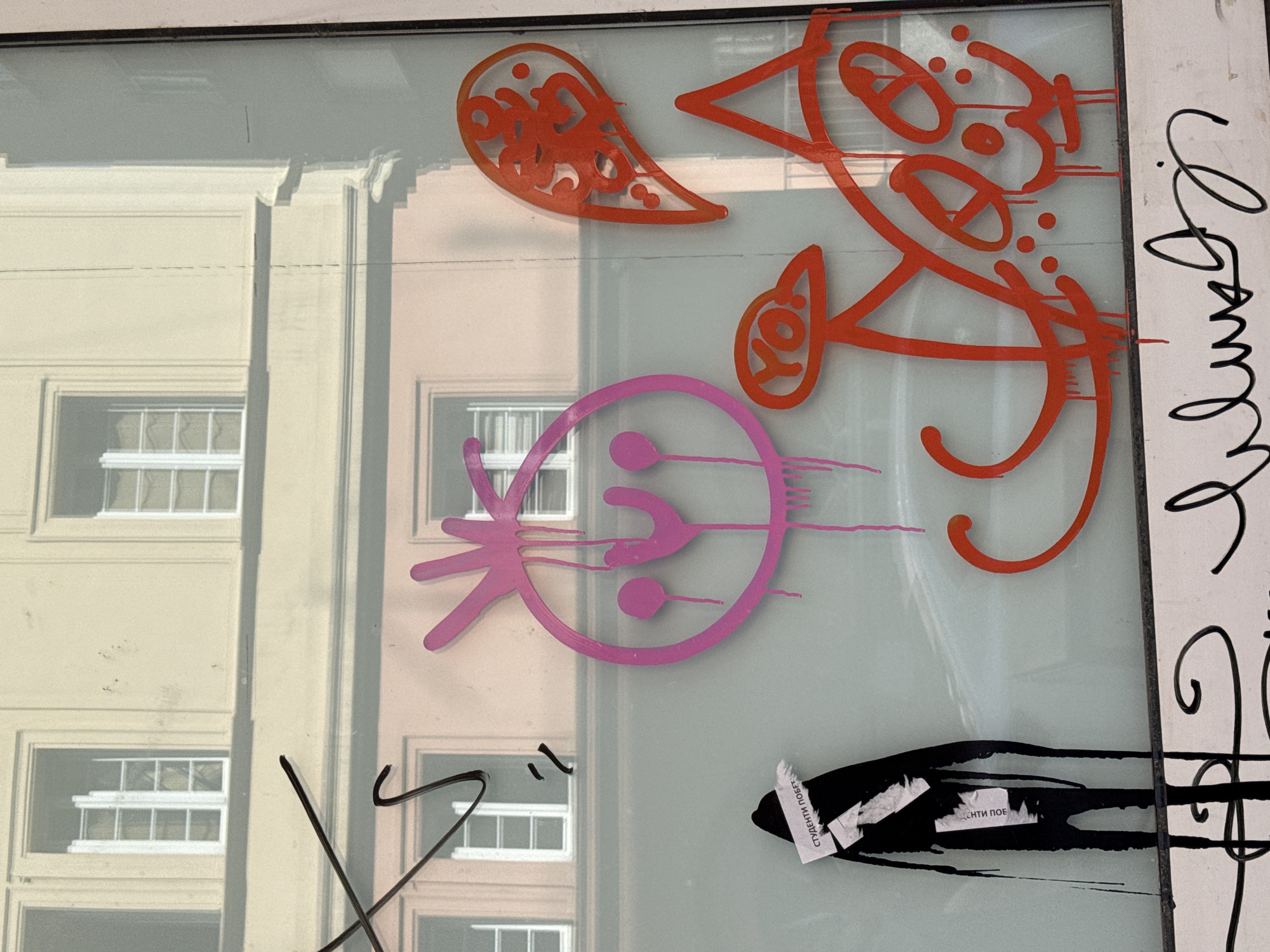 The image shows a window in Belgrade, Serbia, covered with graffiti and stickers. The graffiti is in red, pink, and black, and includes abstract shapes and writing. The sticker is a small, white piece of paper with black writing on it. The window reflects the buildings across the street, showing their windows. The image is taken from a close-up perspective, focusing on the window and its immediate surroundings.