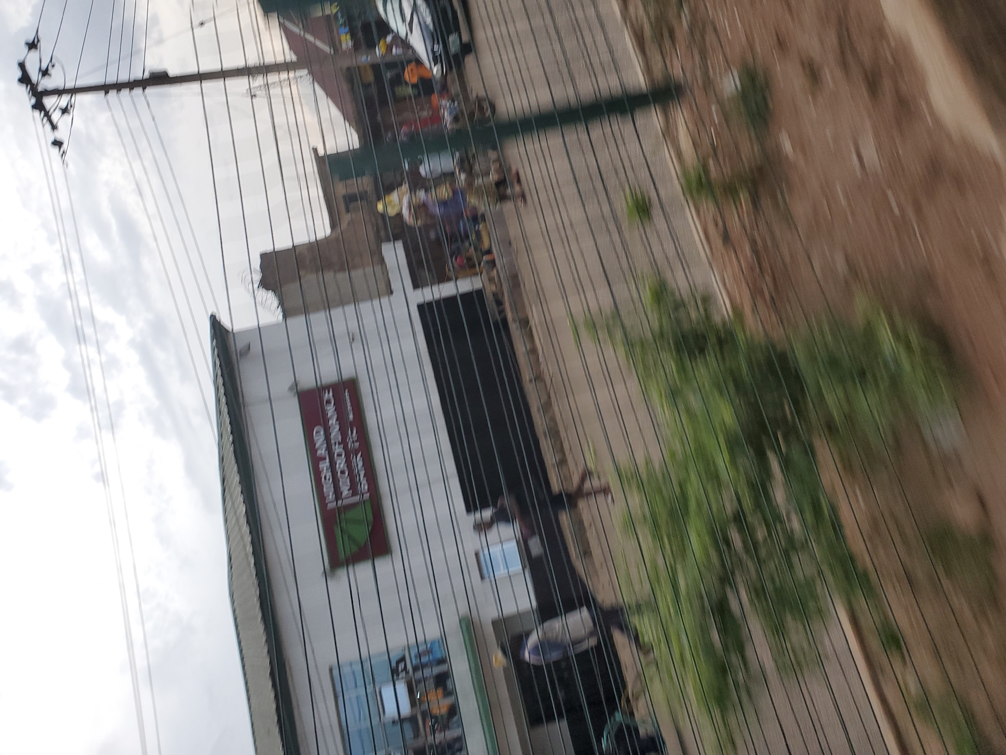 The image depicts a street scene in Bukuru, Nigeria, with a building labeled "HIGHLAND MICROFINANCE BANK PLC." The building has large glass windows and a white facade. Several people are visible walking on the sidewalk or street in front of the building. One person is wearing a blue and white shirt and dark pants, another is wearing a dark shirt and pants, and others are partially obscured. There are also some market stalls or vendor setups with various goods displayed. A black car is parked on the right side of the image. Overhead, there are power lines and a utility pole. The foreground is blurred, suggesting the photo was taken from a moving vehicle, with a wire fence running horizontally across the frame. The sky is overcast.