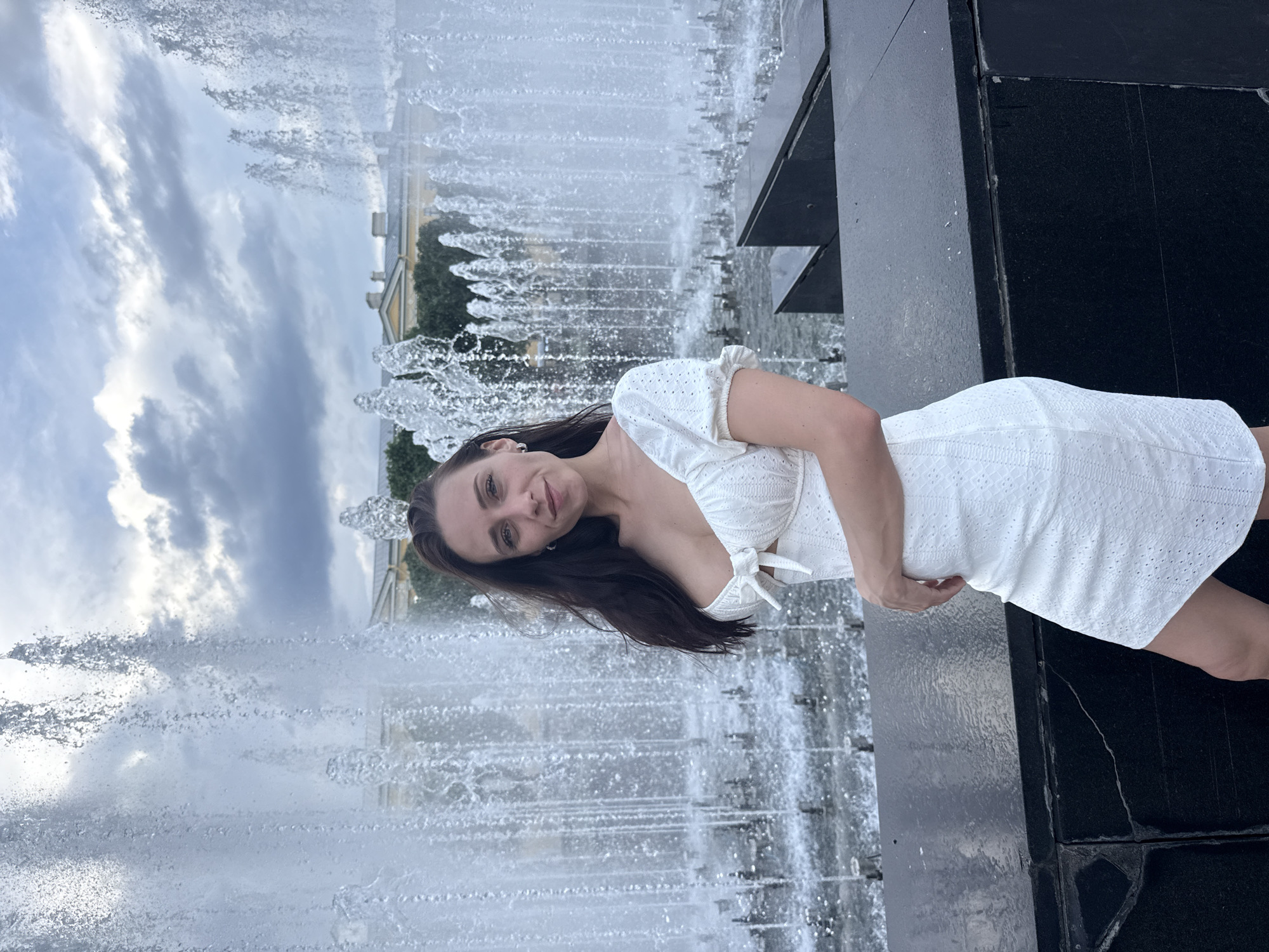 A young woman stands in front of a large fountain with multiple streams of water. She is wearing a white dress with puff sleeves and a bow detail at the chest. The fountain is located in an urban setting, with a building visible in the background, partially obscured by the water spray. The sky is cloudy. The image appears to be taken at the Fountain of Friendship of Peoples in Moscow, Russia, though it is not possible to confirm the exact location from the image alone.