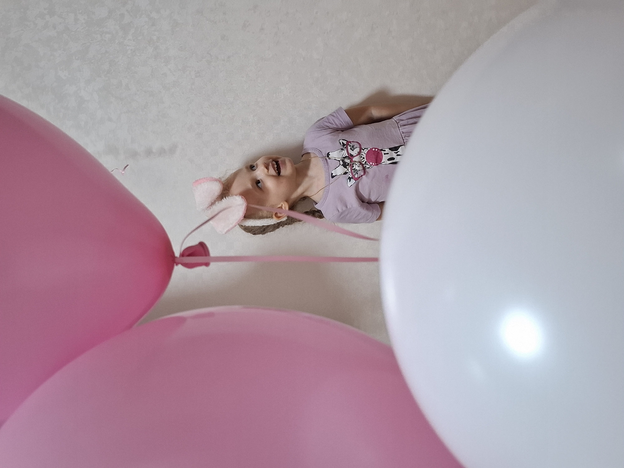 A young girl wearing pink bunny ears and a lavender dress with a giraffe print is lying on a light-colored surface (possibly a bed or floor). Her head is turned towards the camera, and she has an open-mouthed expression. Three balloons are visible: two pink ones are in the foreground, and a larger white one is on the right side of the frame, partially obscuring the girl. The background is a light-colored wall with a subtle pattern. The image was taken in the city of Chaikovsky, Russia.