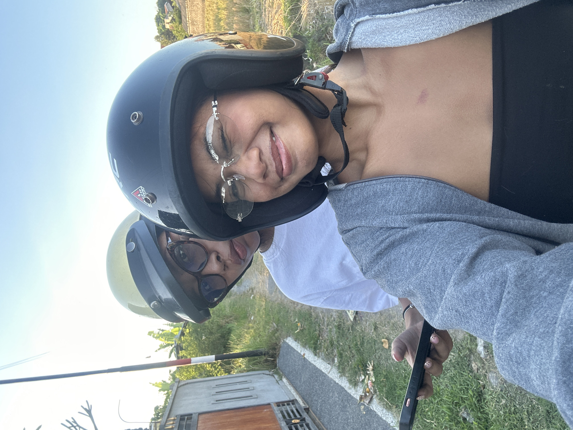 Two individuals, likely a couple, are taking a selfie while riding a motorcycle in Sukawati, Indonesia. The person in the foreground, a young woman with glasses, wears a black helmet and a grey zip-up hoodie over a black tube top. She is smiling broadly, with one eye closed. A small red mark is visible on her chest. Her phone is visible in her left hand. Behind her, a second person, wearing a light yellow helmet and glasses, is looking towards the camera with a neutral expression. They are wearing a white t-shirt. The background shows a rural or suburban setting with a paved road, grass, a fence, trees, and some residential buildings. The sun is setting, casting a warm glow on the scene.