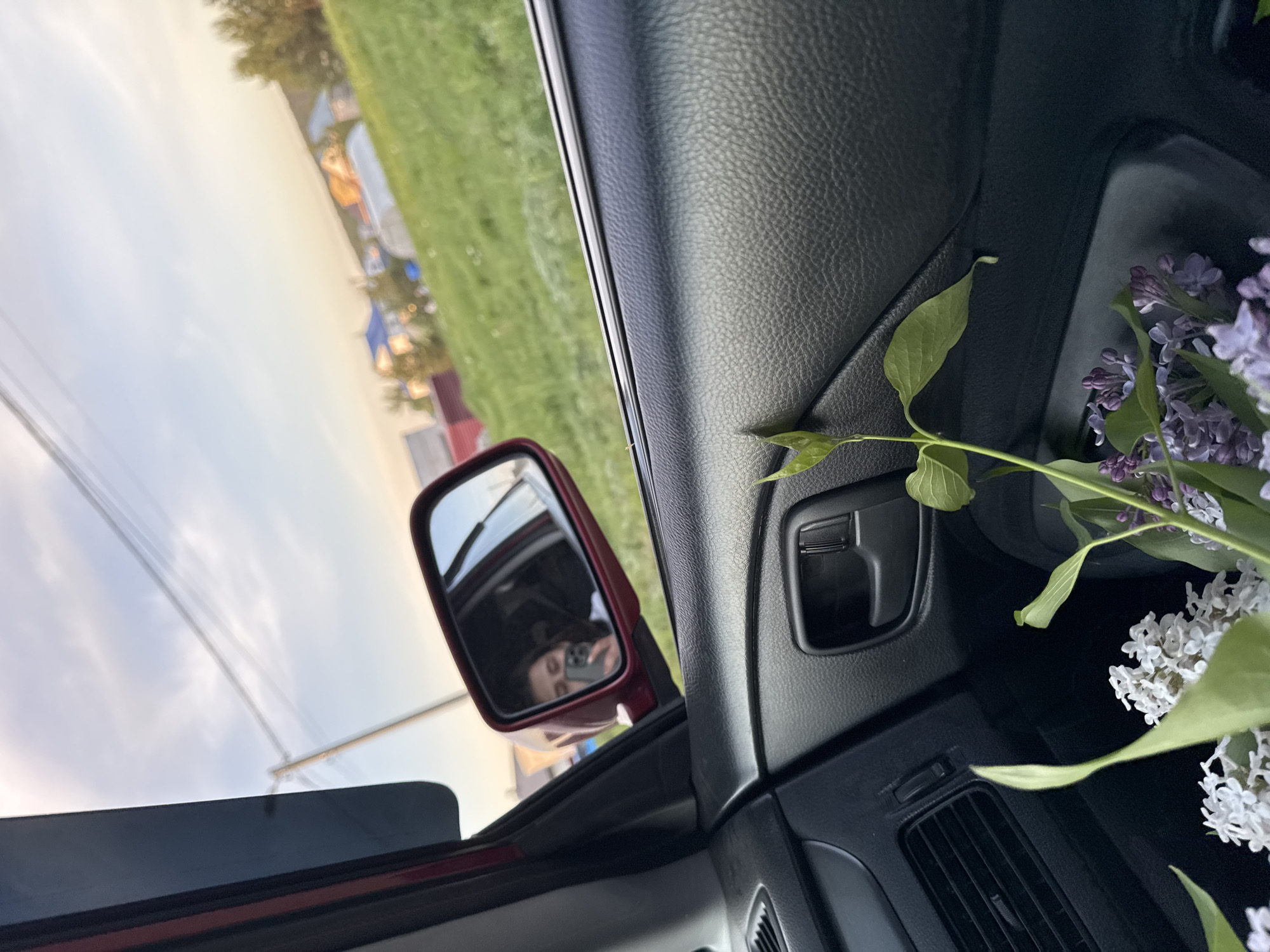 A view from inside a car, showing a side mirror reflecting a person holding a phone. The car is parked or moving slowly, as a green field and buildings are visible through the side window. Lilac flowers are present on the dashboard of the car. The image lacks specific details to conclusively place it in Chaikovsky, Russia, or establish any specific action.
