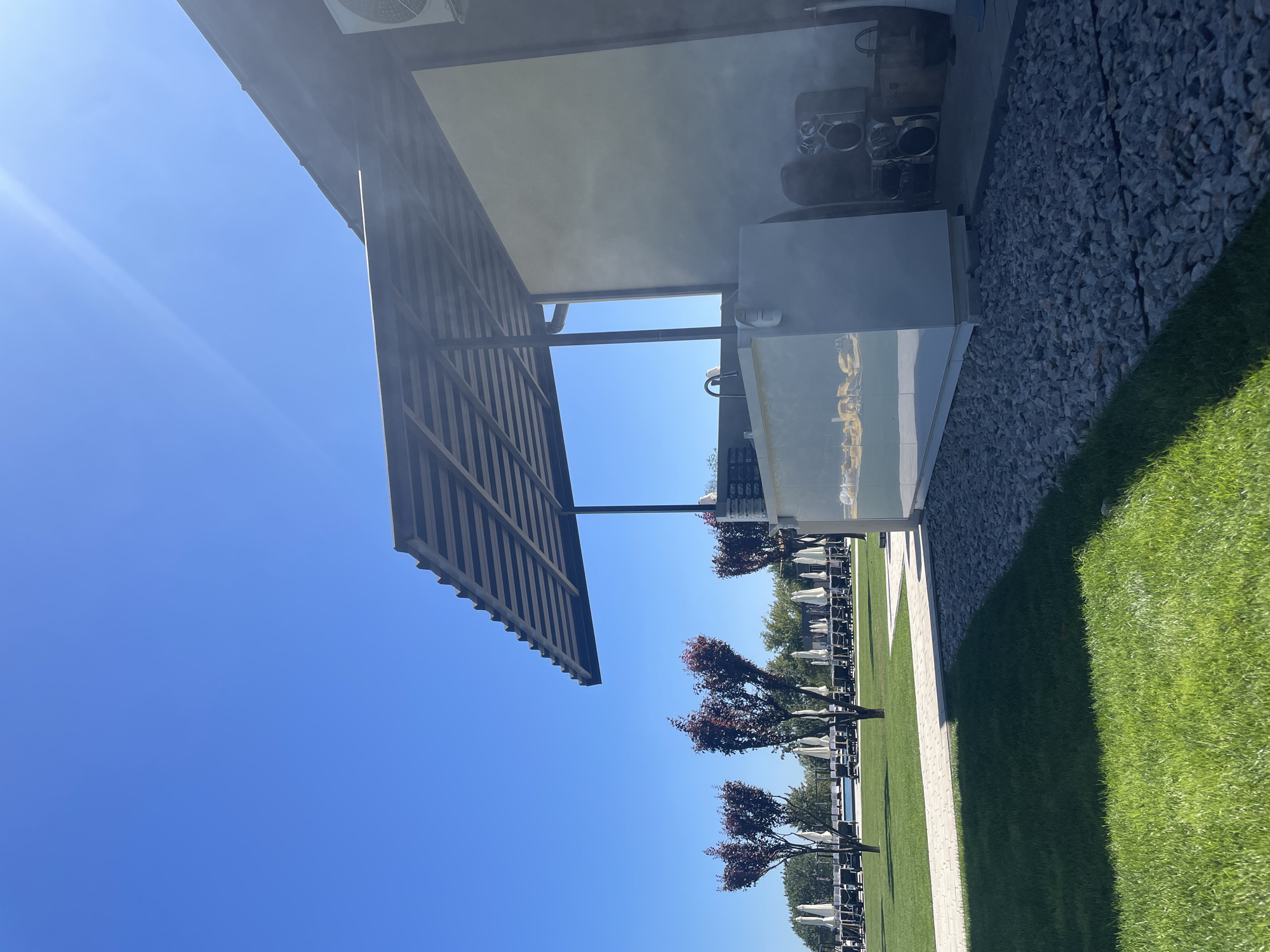 The image captures a sunny outdoor scene at what appears to be a resort or a poolside establishment. In the foreground, a neatly manicured green lawn transitions to a pathway of light-colored gravel. Behind the gravel, a modern, white structure with a metallic awning extends overhead. This structure seems to be a service counter or bar, with a sink visible. To the right of the structure, a boombox and other items are placed on the ground.
In the mid-ground, a row of decorative trees with dark red foliage stands out against a backdrop of loungers and white umbrellas, suggesting a seating area for guests. A swimming pool is partially visible between the trees and loungers. The background consists of more trees and a clear, bright blue sky with a sunbeam creating a lens flare. The overall environment is clean, organized, and set for outdoor leisure.