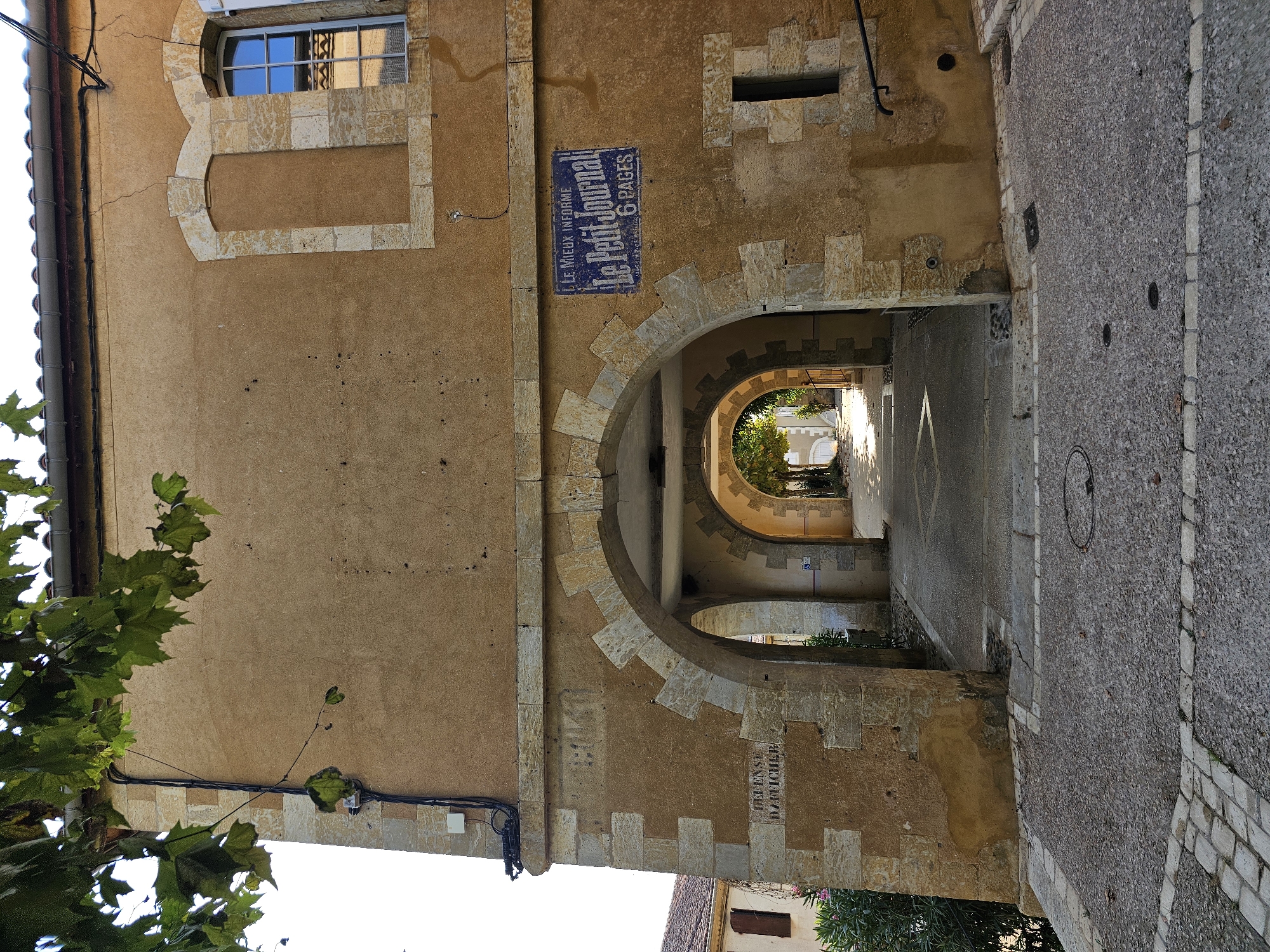 The image shows an arched passage through a building in Lavardens, France. The building has a stucco facade, arched openings, and stone detailing. Above the right arch, a blue sign reads "LE MIEUX INFORME Le Petit Journal 6 PAGES". To the left of the arches, a sign reads "DEFENSE D'AFICHIR". The passage leads to a sunlit street lined with buildings. The foreground is paved with cobblestones and gravel. Green foliage is visible in the upper left corner. The scene appears to be a historical town square or thoroughfare.