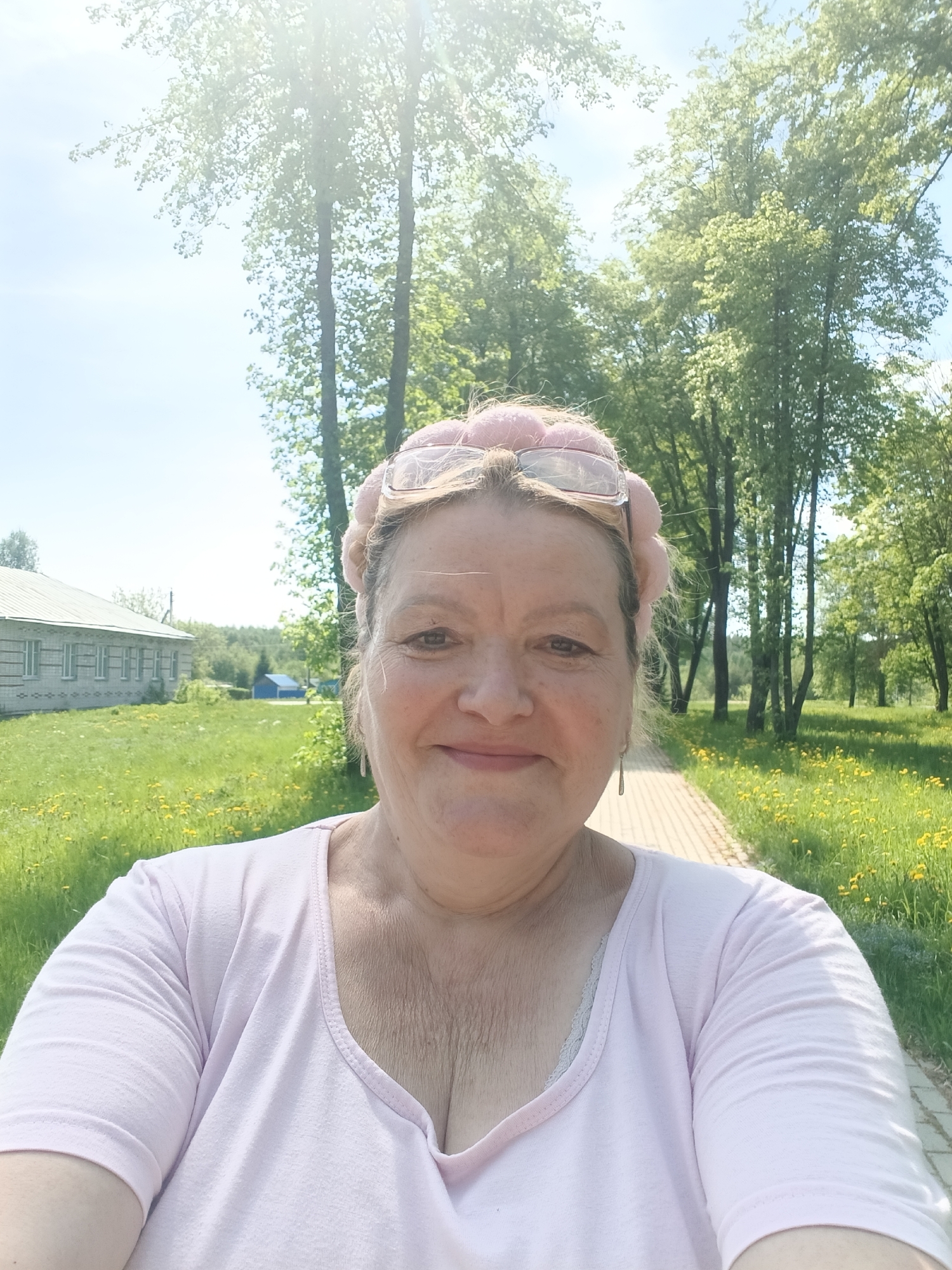 A selfie photograph shows a woman outdoors in Красный Маяк, Russia. She is smiling and wearing a pink shirt, a fuzzy pink headband with glasses attached, and dangle earrings. A brick path leads away from her into a grassy area with yellow wildflowers. Trees are visible on both sides of the path and behind the woman. To the left is a building with multiple windows. The sky is blue with some clouds and sunlight.