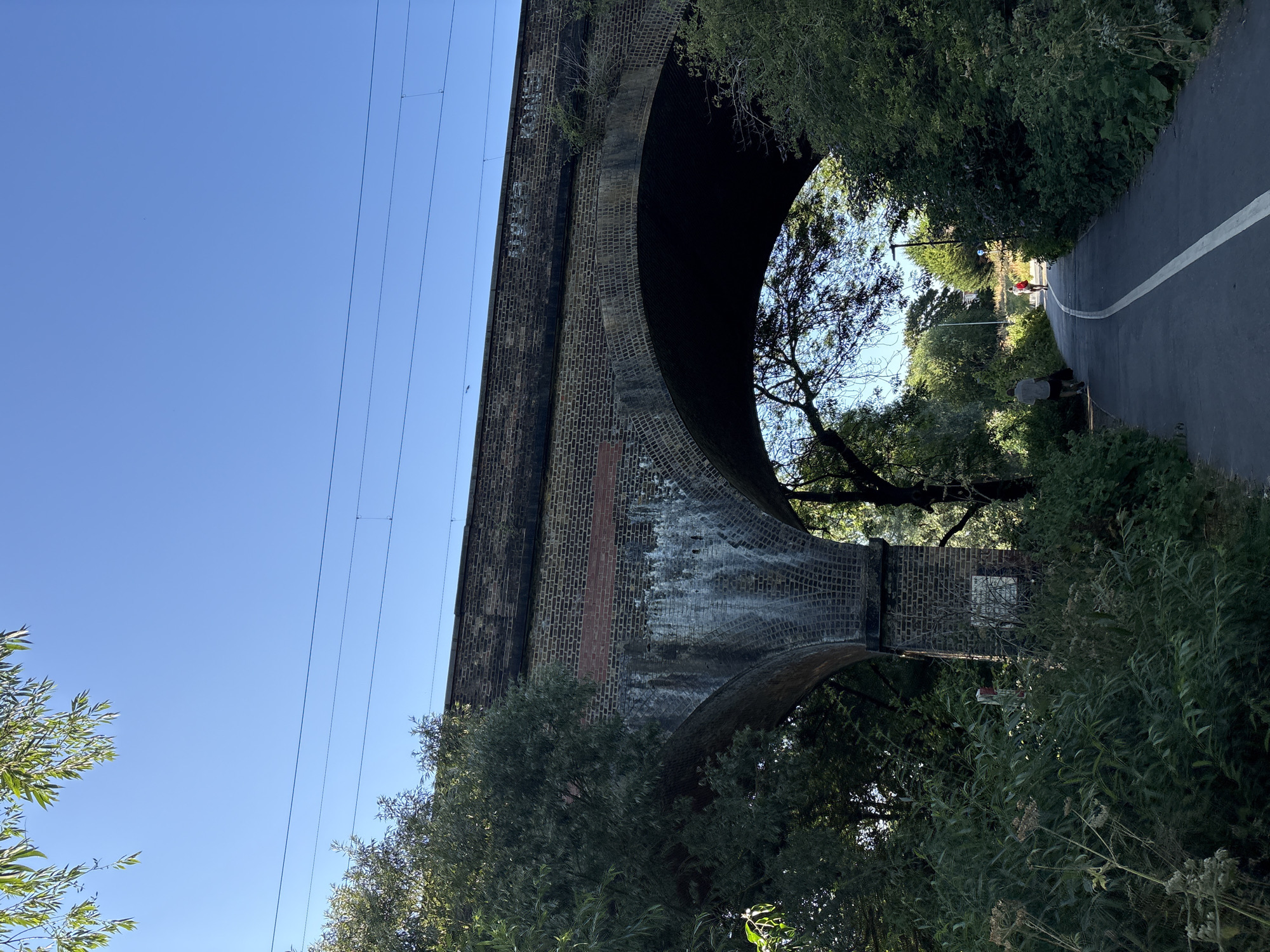 A paved pathway, marked with a white line, curves through lush green foliage under a clear blue sky. Two individuals are visible on the path, one further ahead wearing a red backpack and the other closer to the viewer, facing away. Overhead, a large brick railway viaduct with arched openings dominates the scene, its upper edge lined with railway tracks and overhead wires. The viaduct shows signs of age with some discoloration and graffiti. The surrounding environment is densely vegetated, with trees and plants framing the pathway and the viaduct. The overall scene suggests a public path in a semi-urban or parkland setting, likely part of a larger greenway or recreational area in Chelmsford, United Kingdom, possibly near the River Chelmer or a similar waterway where such viaducts are common.