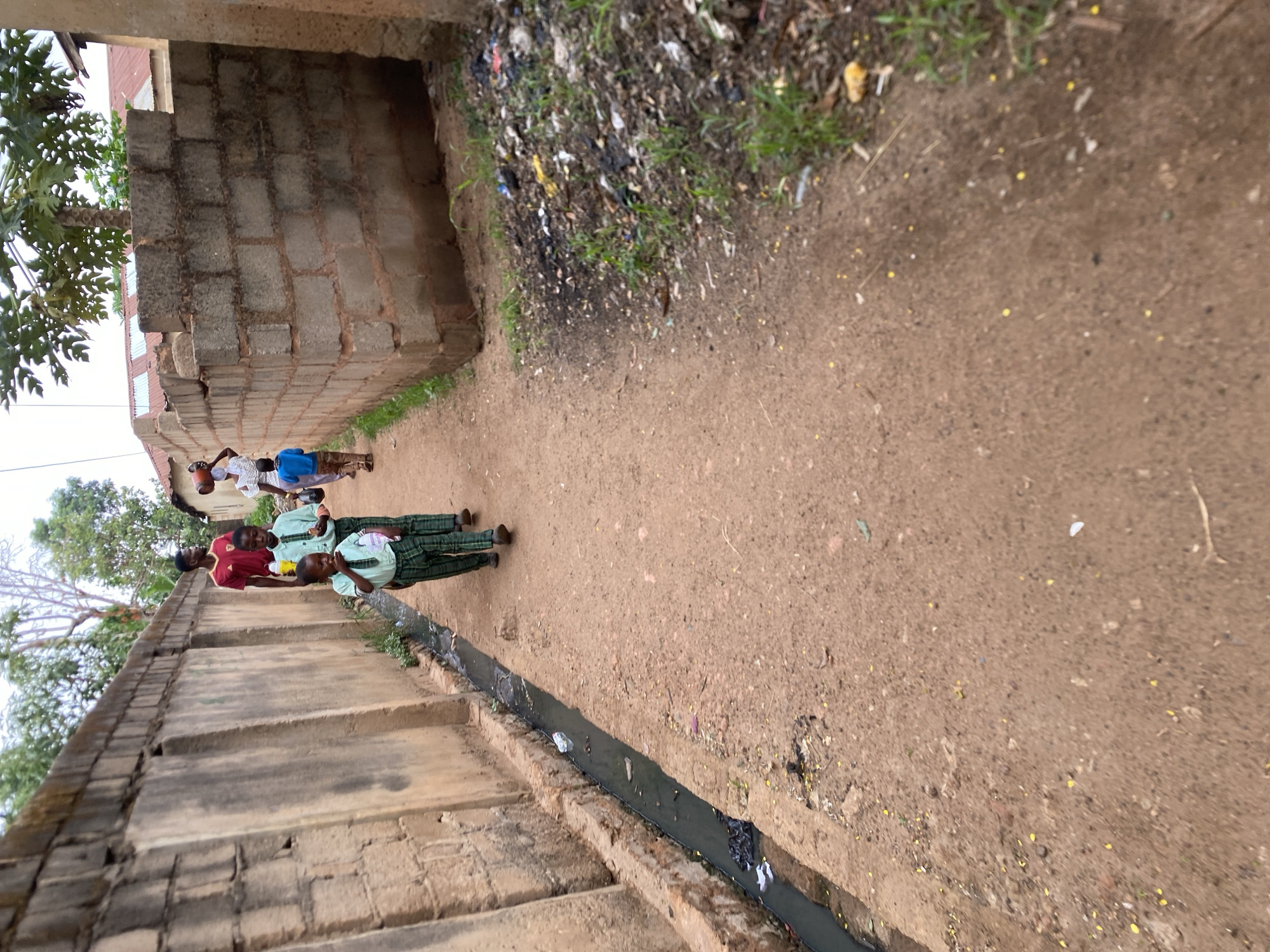 In Bukuru, Nigeria, a narrow dirt path is bordered by a block wall on one side and a debris-strewn area on the other. A drainage ditch with stagnant water runs along the base of the wall. Four individuals, including three children in school uniforms, walk along the path. One man is wearing a red shirt. A tree and house are visible in the background.