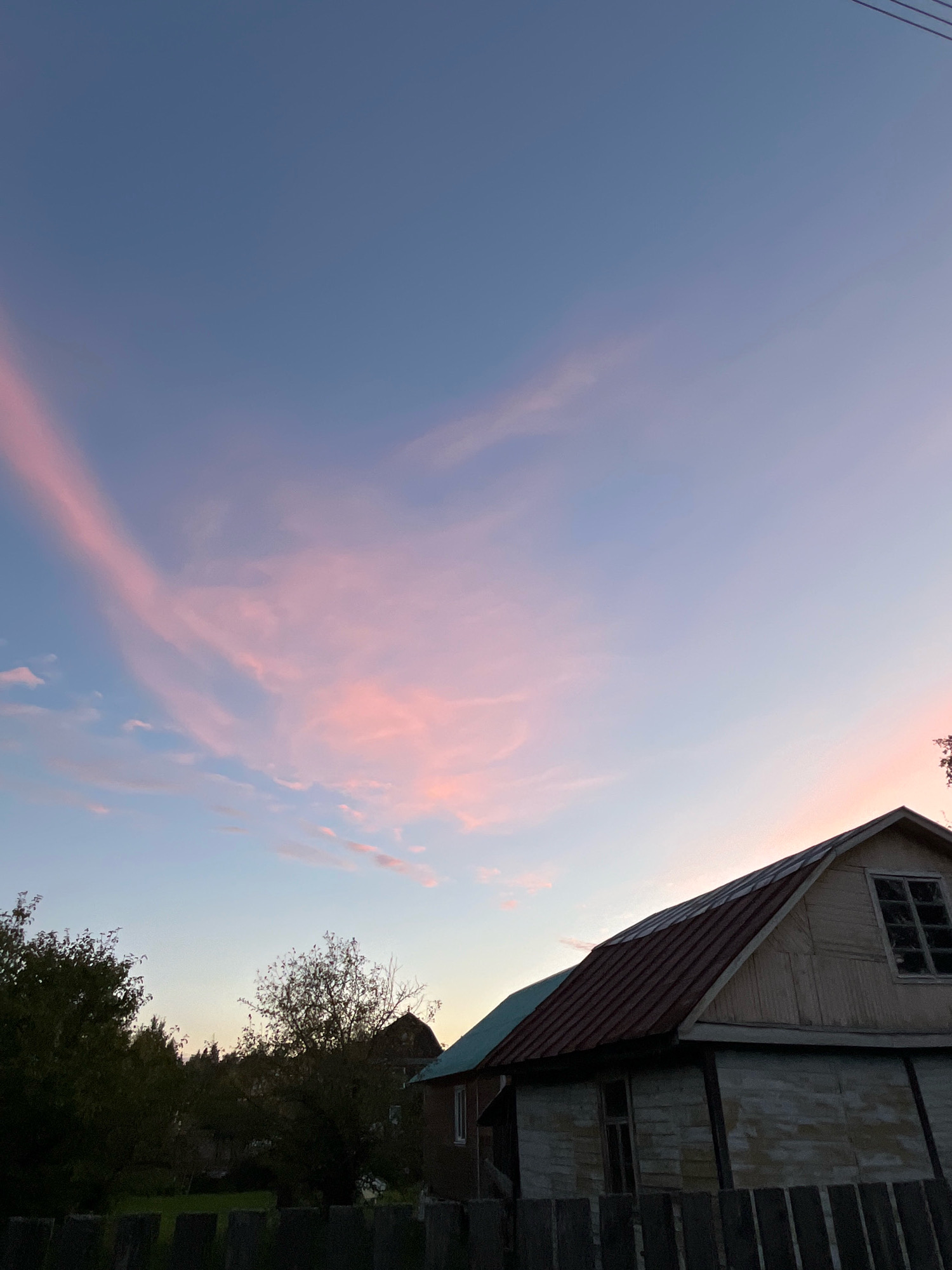 The image depicts a rural scene at dusk, likely in the city of Krasnaya Presnya SNT, Russia. The sky is a gradient of deep blue at the top transitioning to soft pink and lighter blue hues near the horizon, indicative of sunset. Wispy pink clouds are scattered across the upper portion of the sky. In the foreground, the roofline and upper portion of a weathered wooden house with a dark metal roof are visible on the right side. A window with multiple panes is set into the gable end of the house. To the left of the house, trees with dark foliage are silhouetted against the fading light. A wooden fence runs along the very bottom of the image, obscuring the lower part of the scene. The overall atmosphere is tranquil and natural.